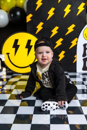 Baby boy in black hoodie sits on checkered floor at lightning bolt smiley face themed first birthday party.