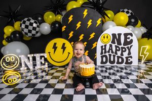 Baby's first birthday cake smash with smiley face balloons, lightning bolts, and One Happy Dude sign on checkered floor.