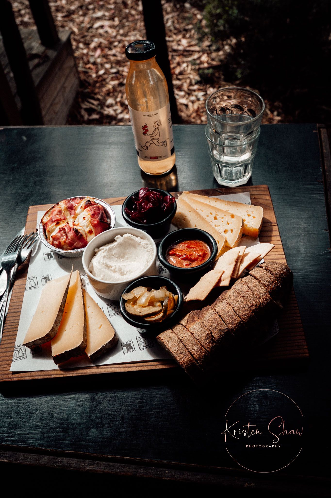 Rustic cheese board with assorted cheeses, crackers, jams, and condiments served with a bottle of white wine on a dark table.