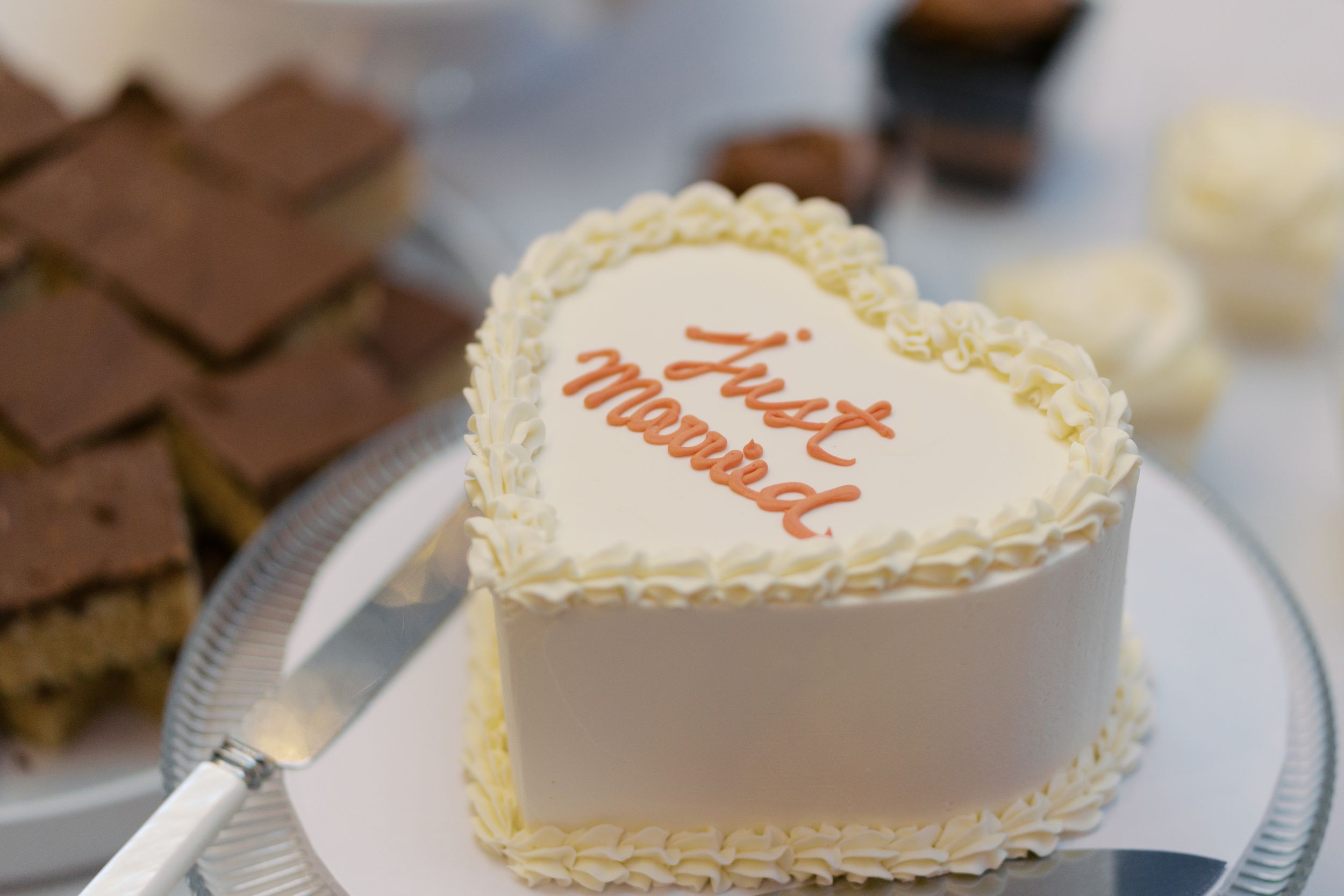 Heart-shaped white wedding cake decorated with 'Just Married' in orange cursive frosting on a silver cake stand.