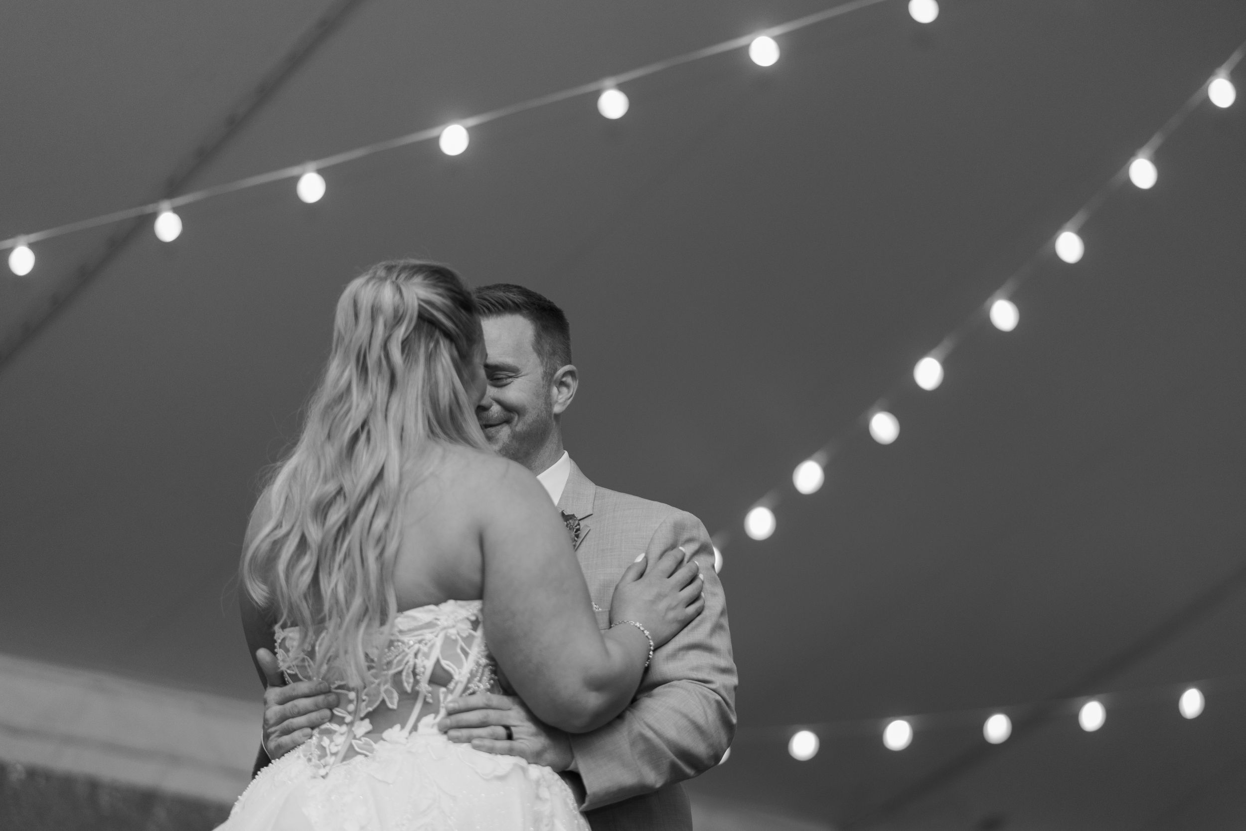 Black and white photo of bride and groom sharing first dance under string lights at elegant wedding reception.