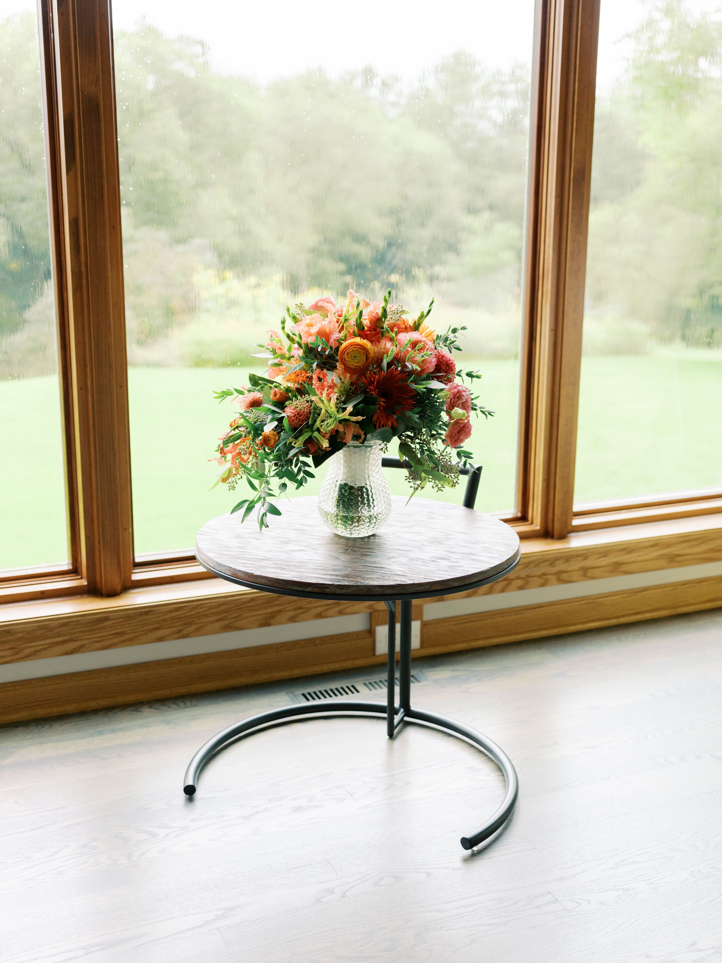 A vibrant floral bouquet in a white vase sits on a round side table by a large window overlooking lush greenery.