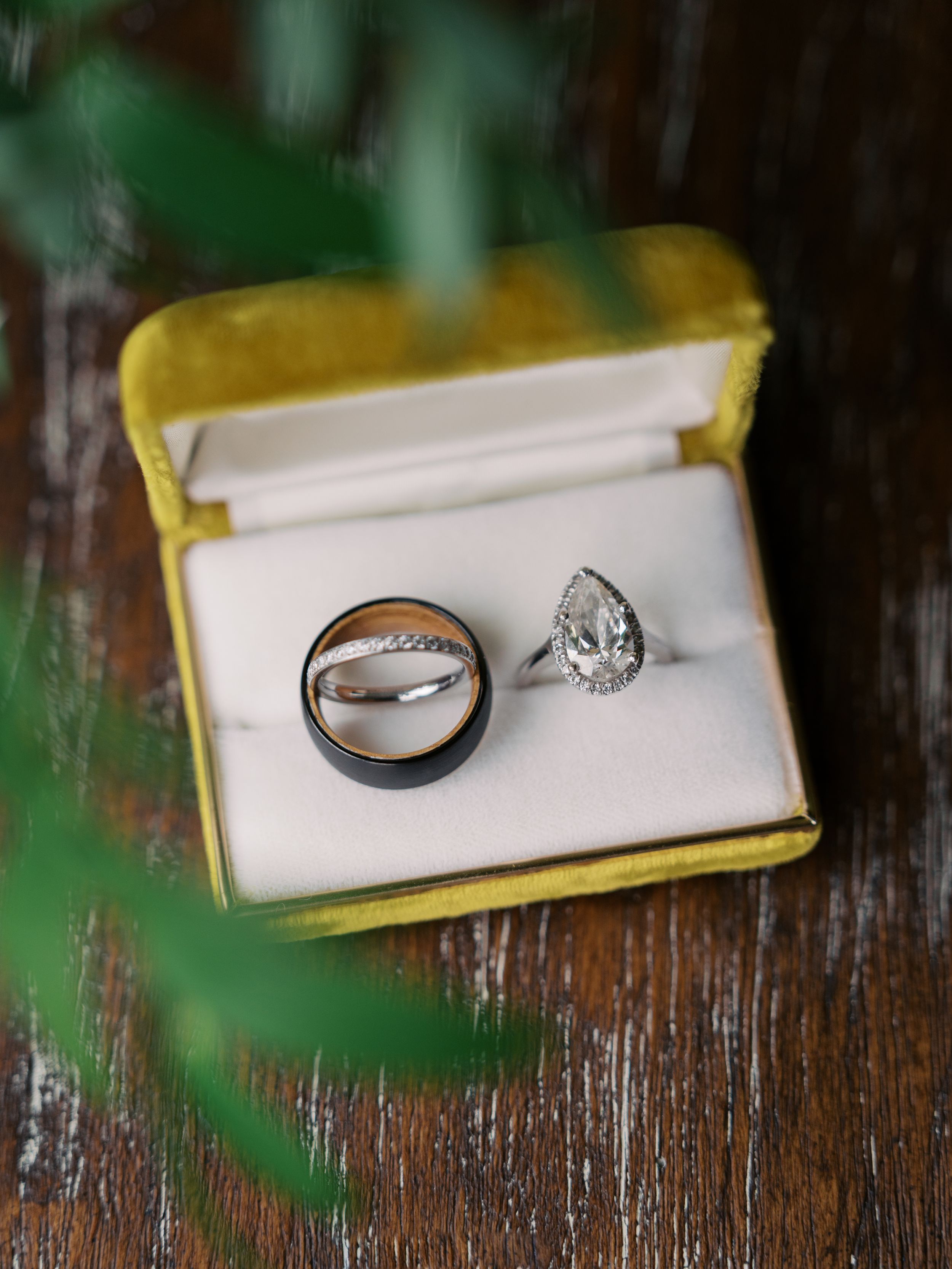 Three wedding rings displayed in a yellow velvet box, including a black band, diamond band, and pear-shaped halo engagement ring.