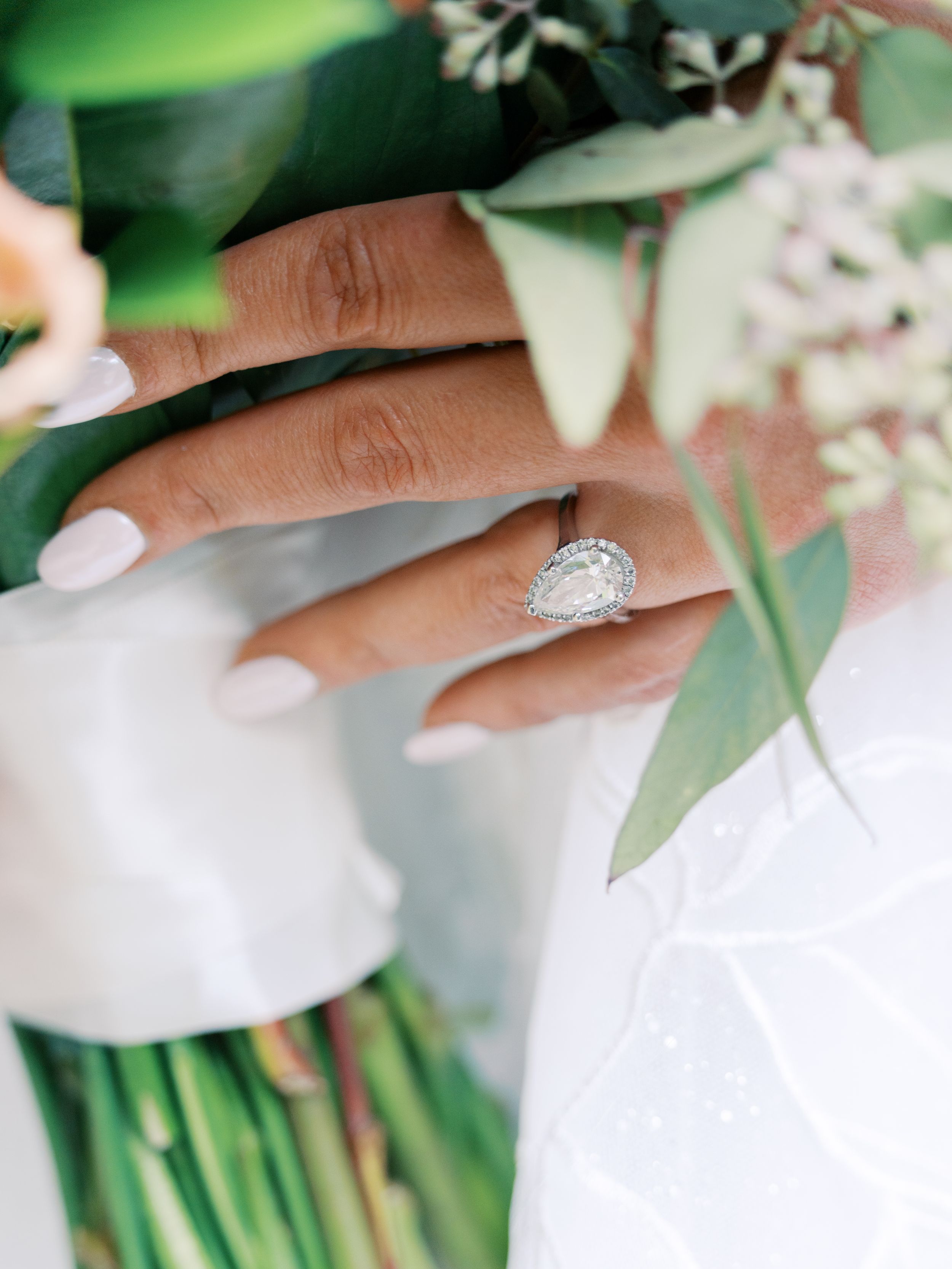 Close-up of a pear-shaped diamond halo engagement ring on a bride's hand holding a floral bouquet.
