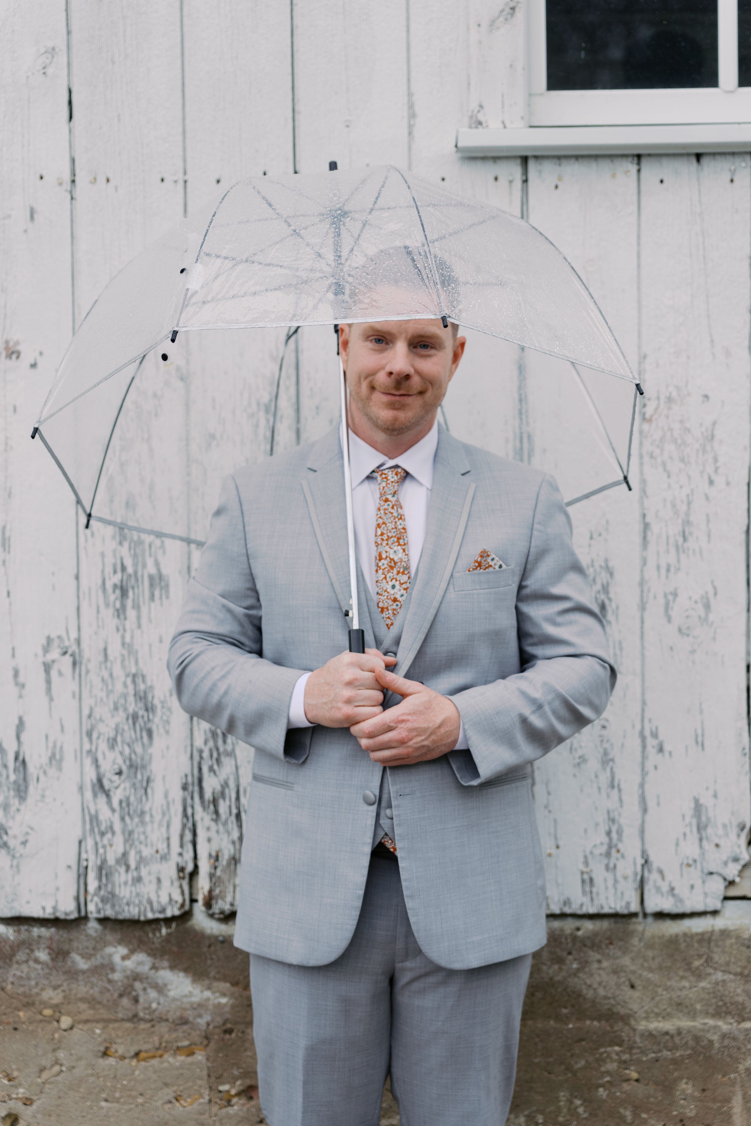 A groom in a light gray suit with floral tie holds a clear umbrella while standing in front of a rustic white barn door.