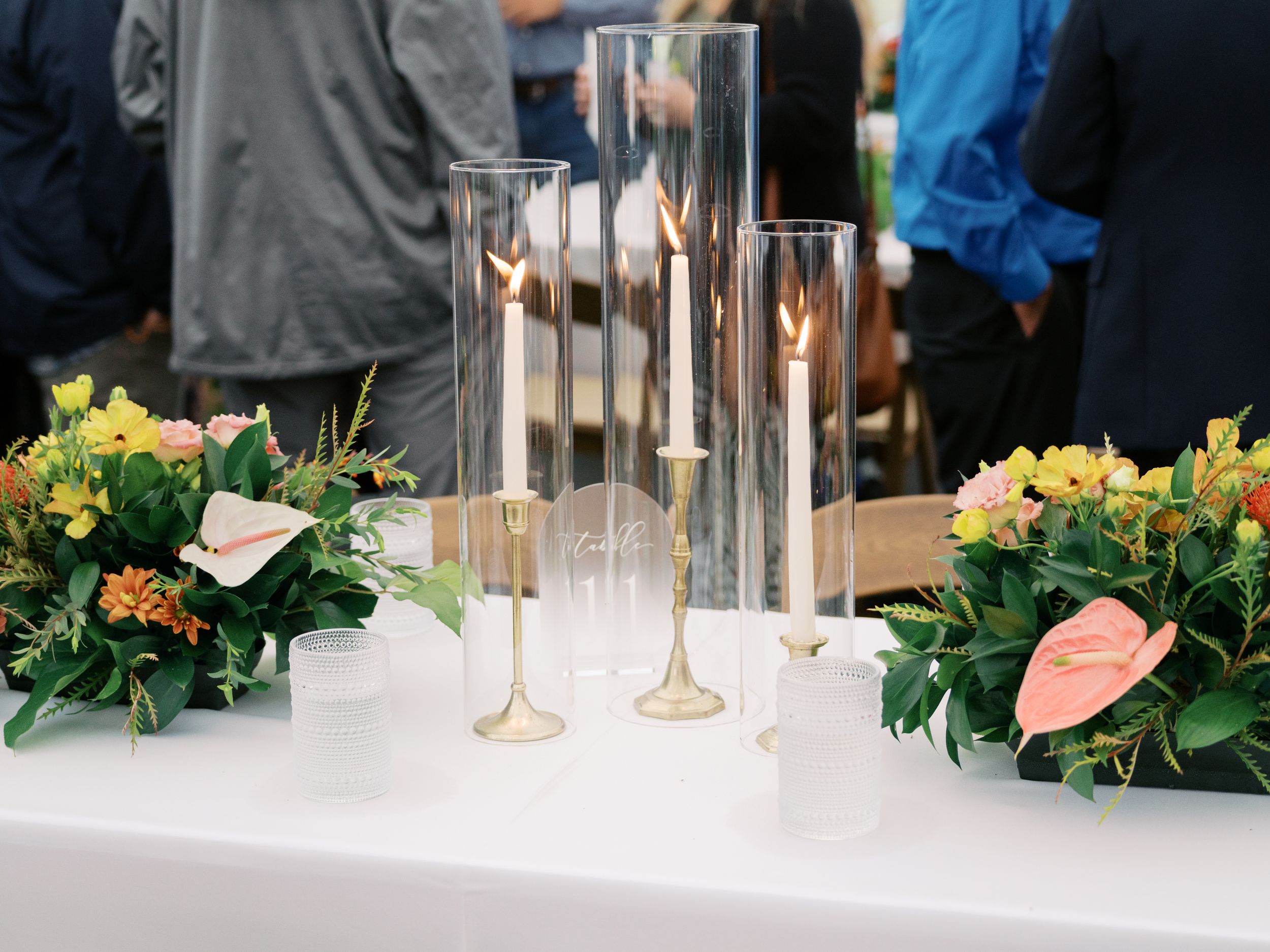 Elegant wedding reception table with tall glass candle holders, gold candlesticks, and floral arrangements with pink anthuriums.