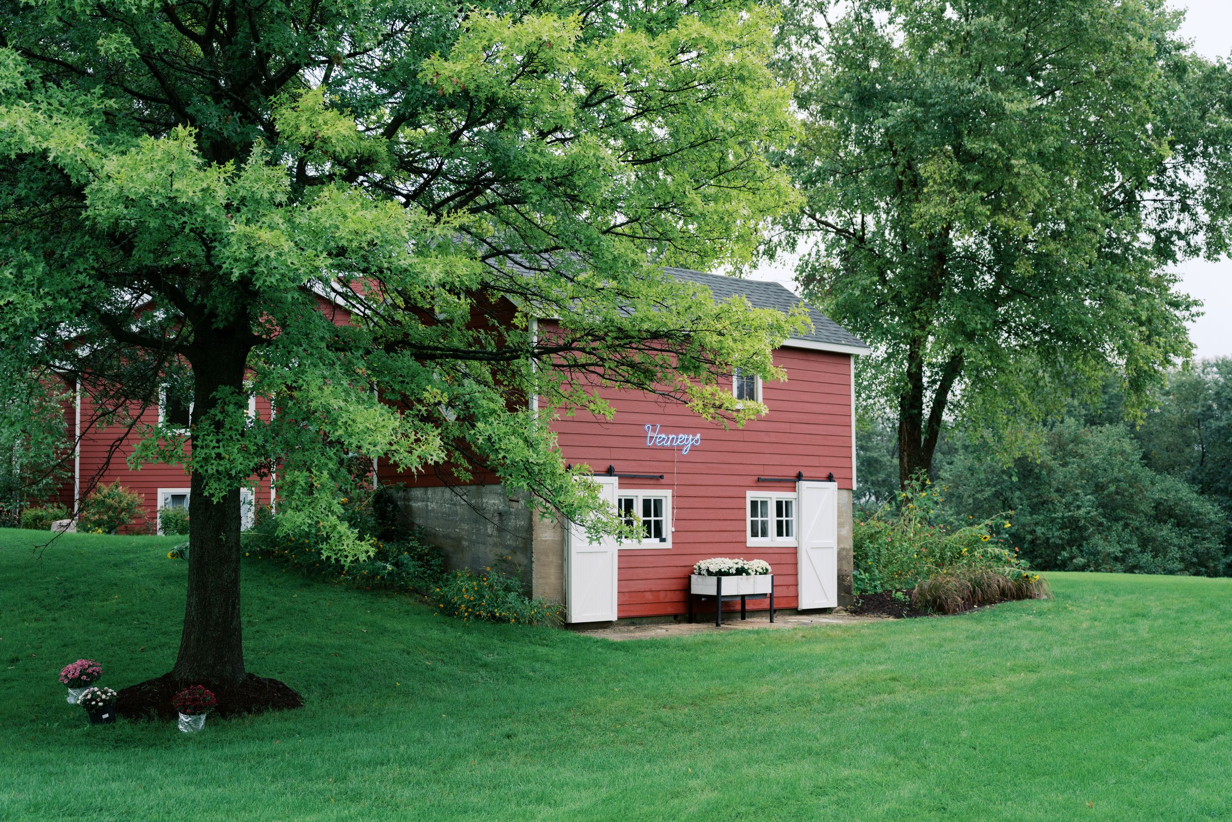 Red barn-style house surrounded by lush green trees and manicured lawn on a cloudy day.
