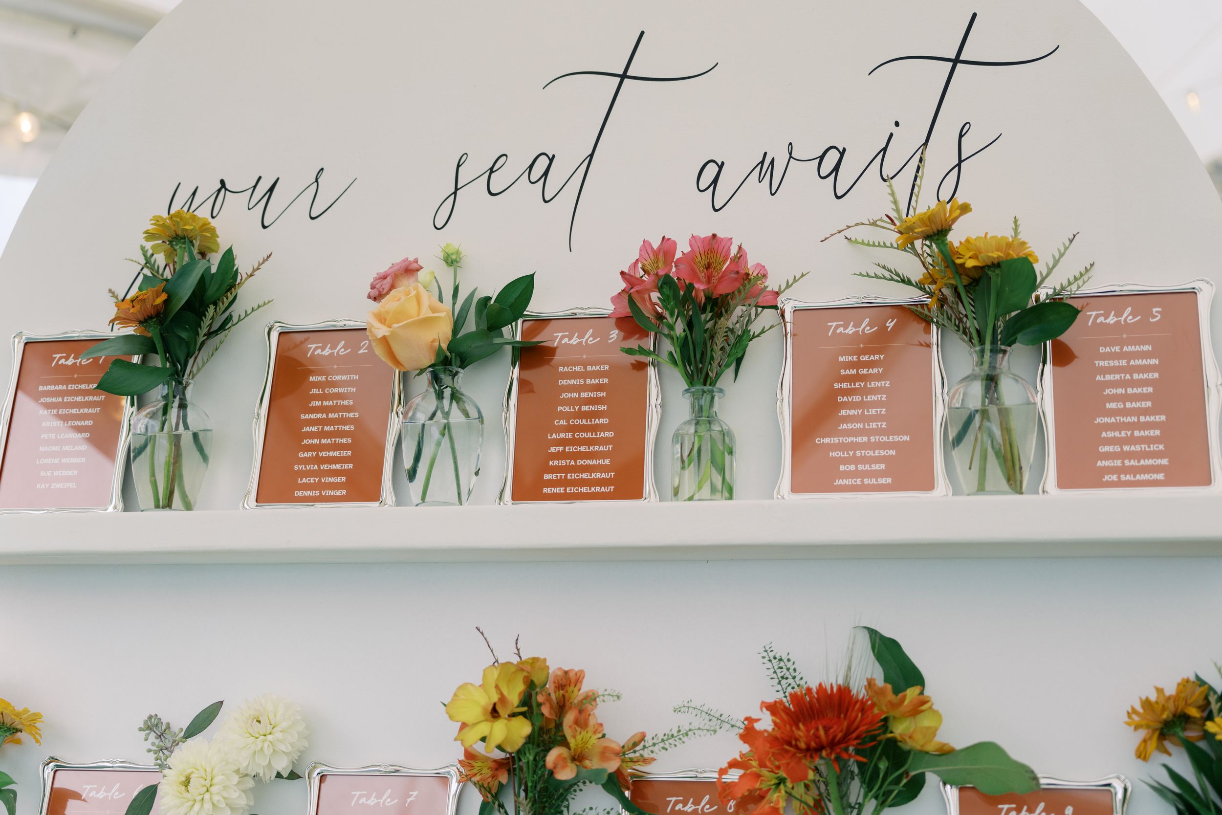 Wedding seating chart display with mason jar floral arrangements and cursive 'your seat awaits' sign on white shelf.