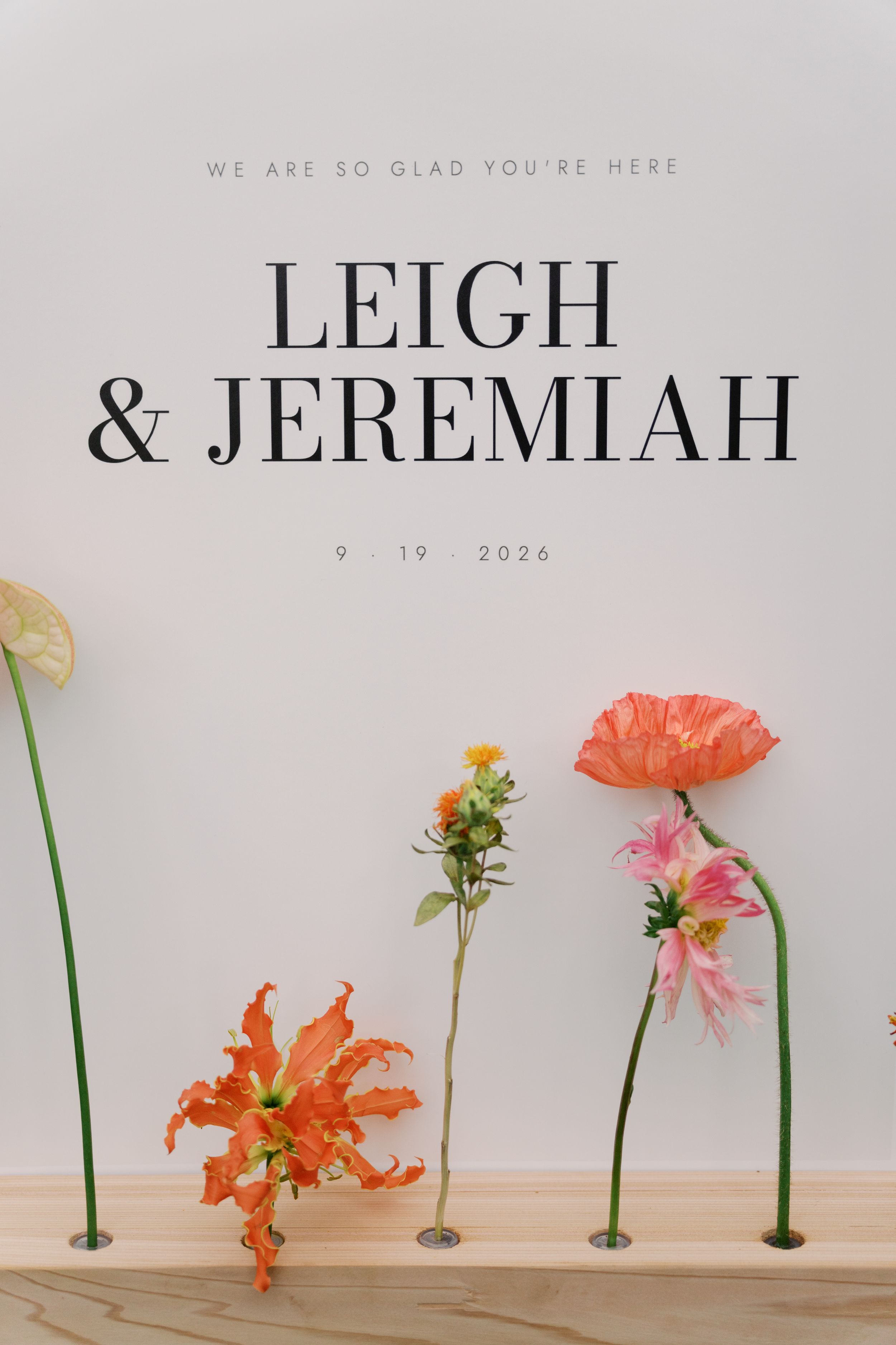 Elegant wedding welcome sign for Leigh and Jeremiah on 9.19.2026 featuring colorful flowers on a wooden shelf.