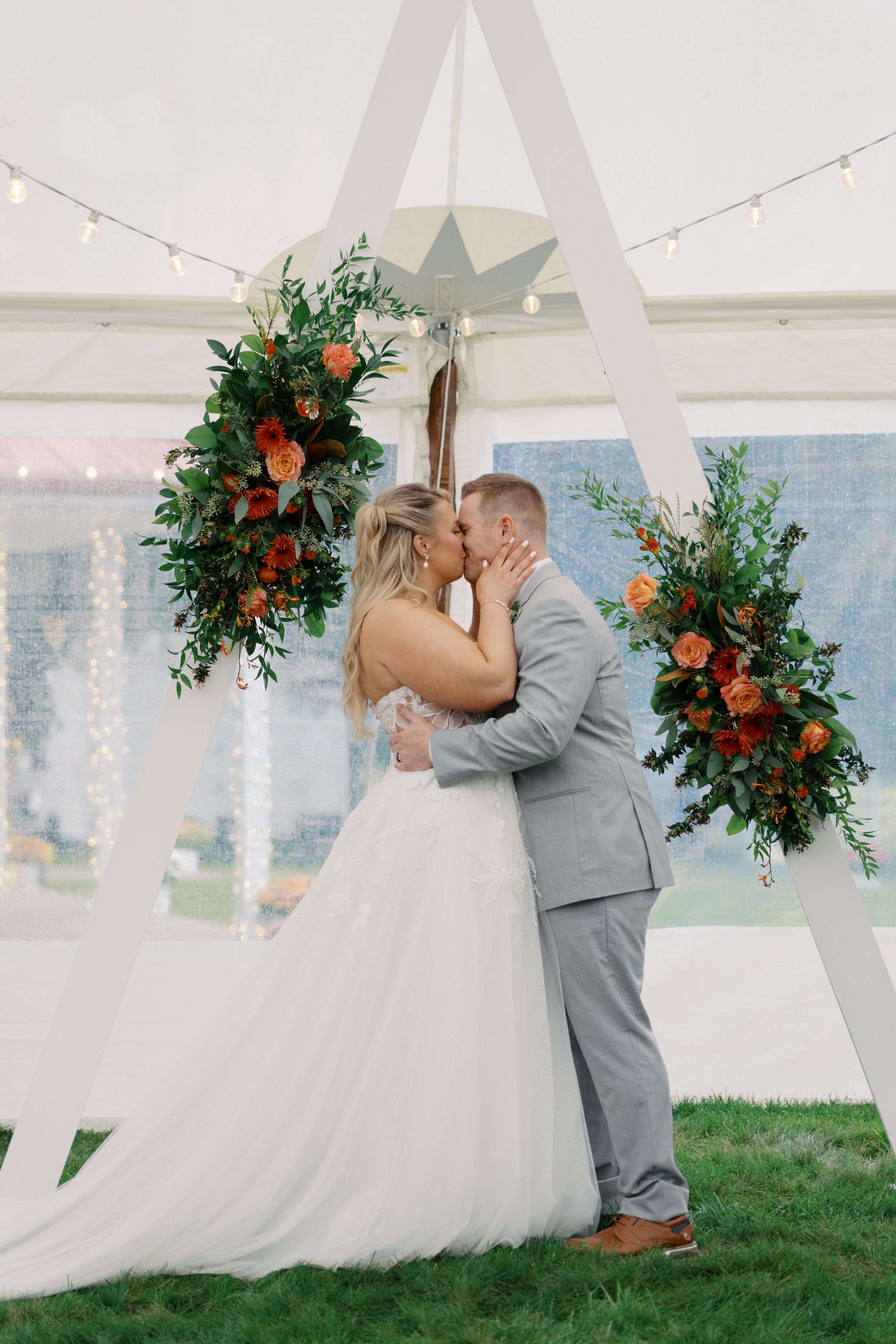 Bride and groom share first kiss under floral arch with orange roses and greenery at outdoor tented wedding ceremony.