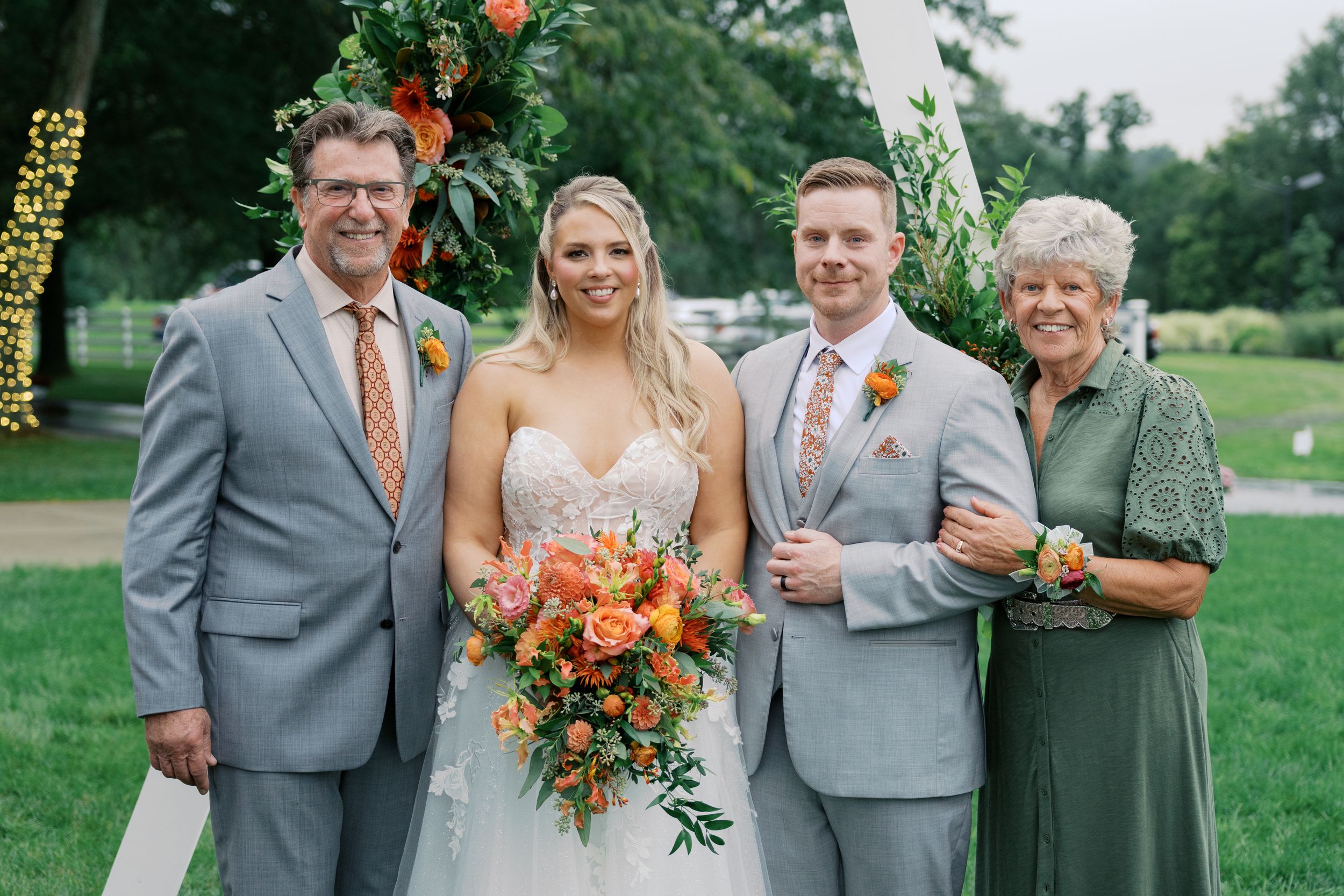 Wedding party of four posing outdoors, bride holding orange bouquet, groom and older man in gray suits, woman in green dress.