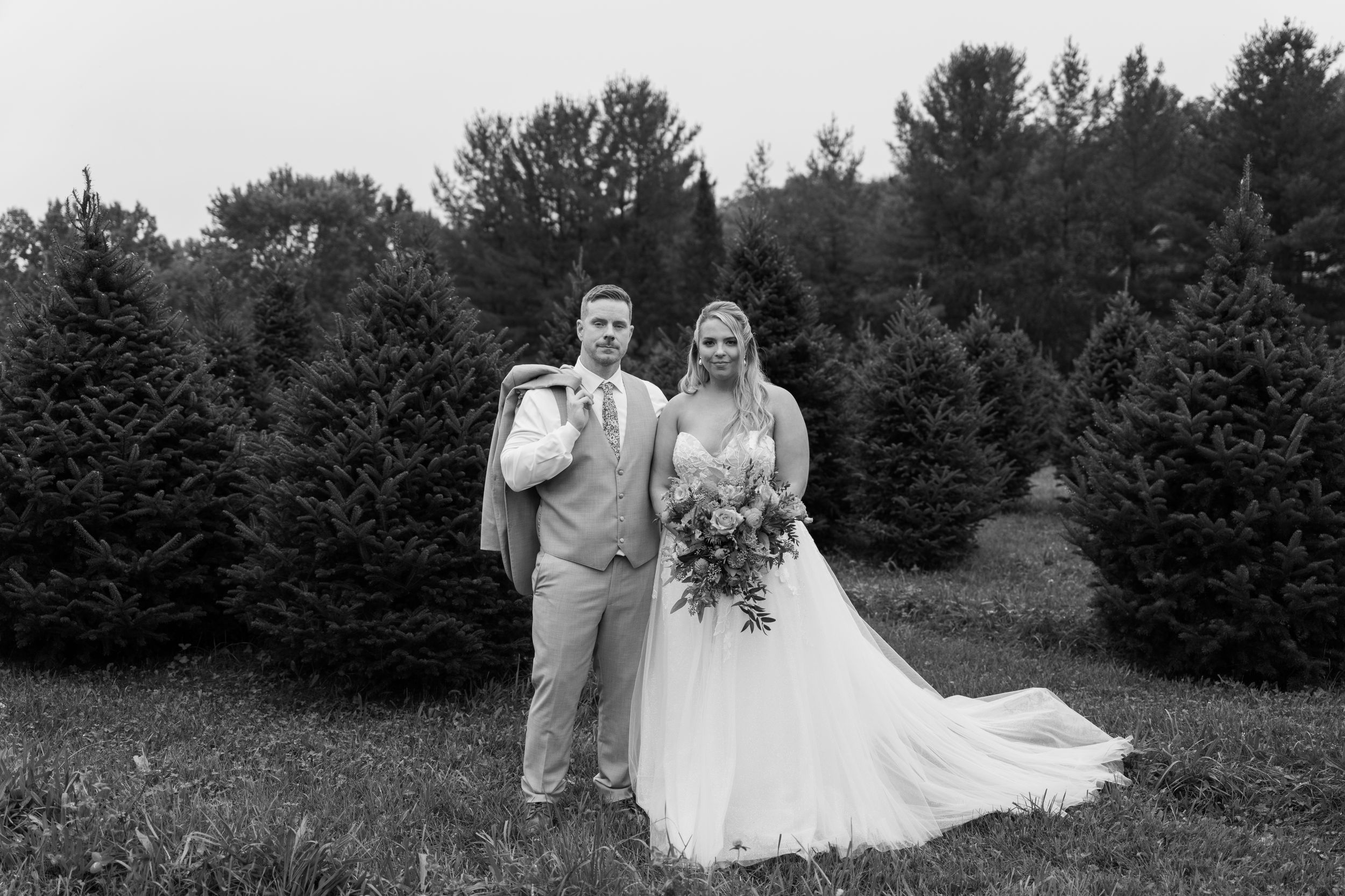 Bride and groom posing in front of evergreen trees in a black and white wedding portrait outdoors.