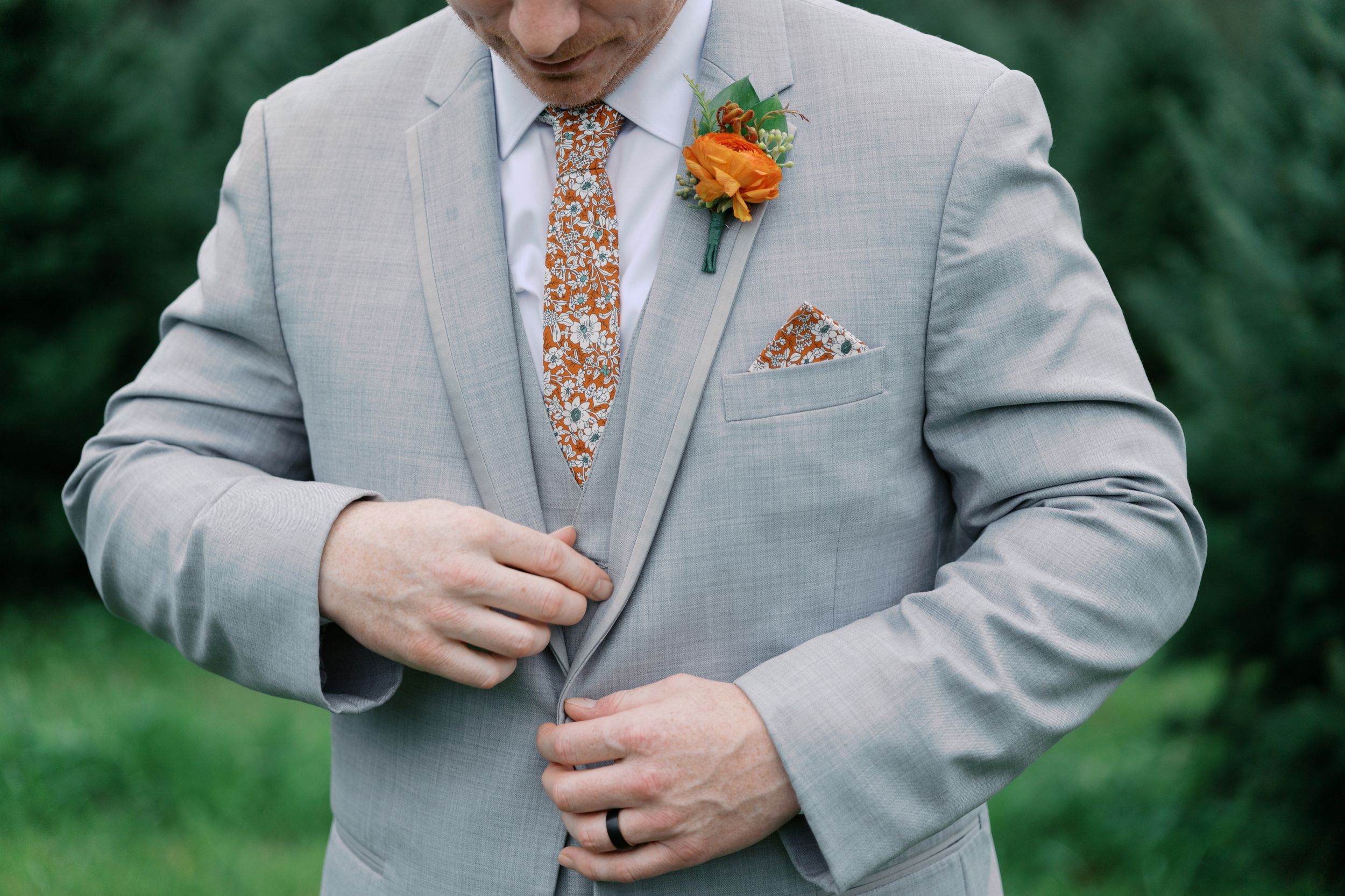 Man in gray suit adjusting jacket, wearing floral orange tie, matching pocket square, and orange rose boutonniere.