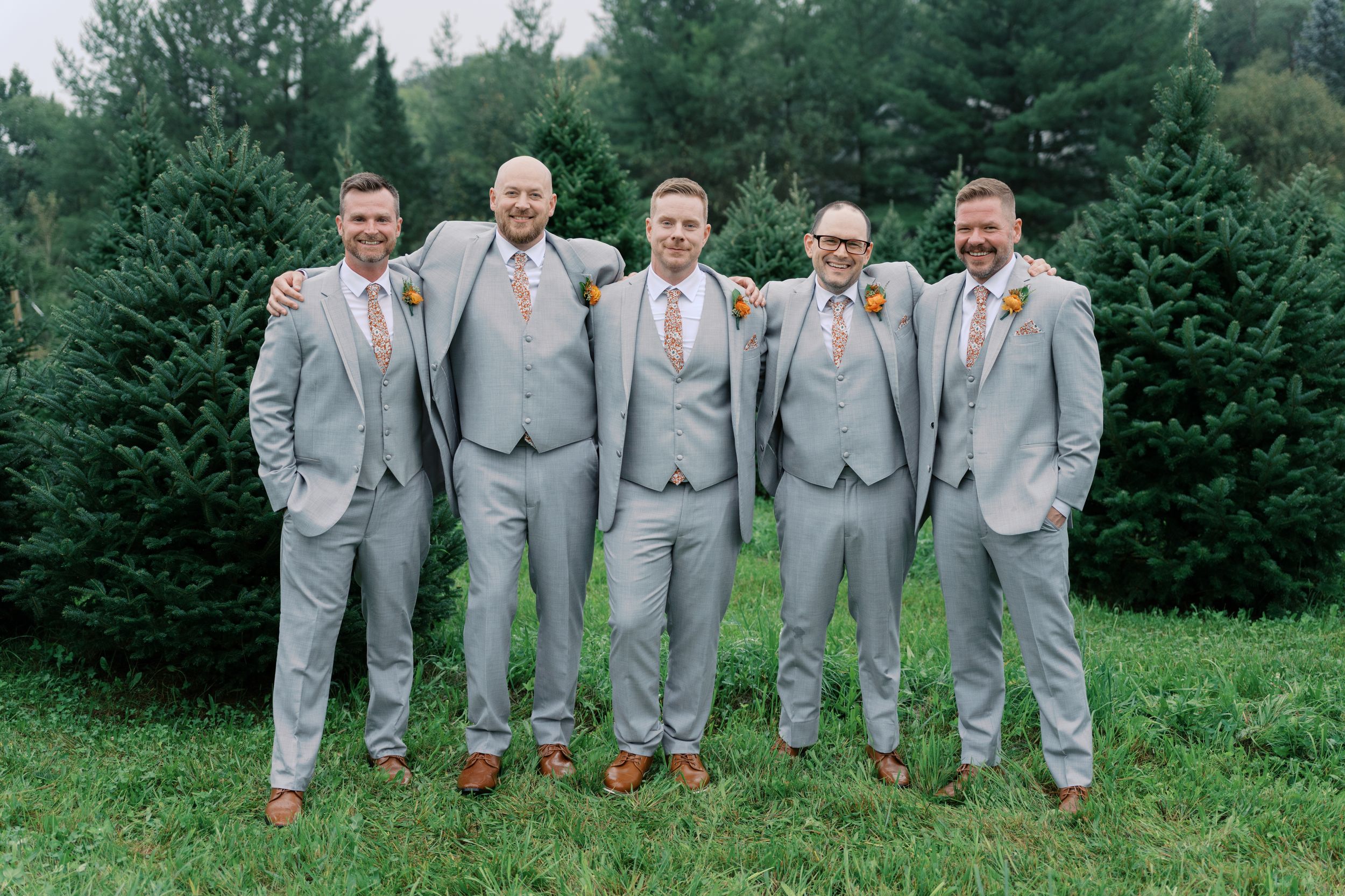 Five groomsmen in matching light gray suits with floral ties pose together on green grass with evergreen trees behind them.