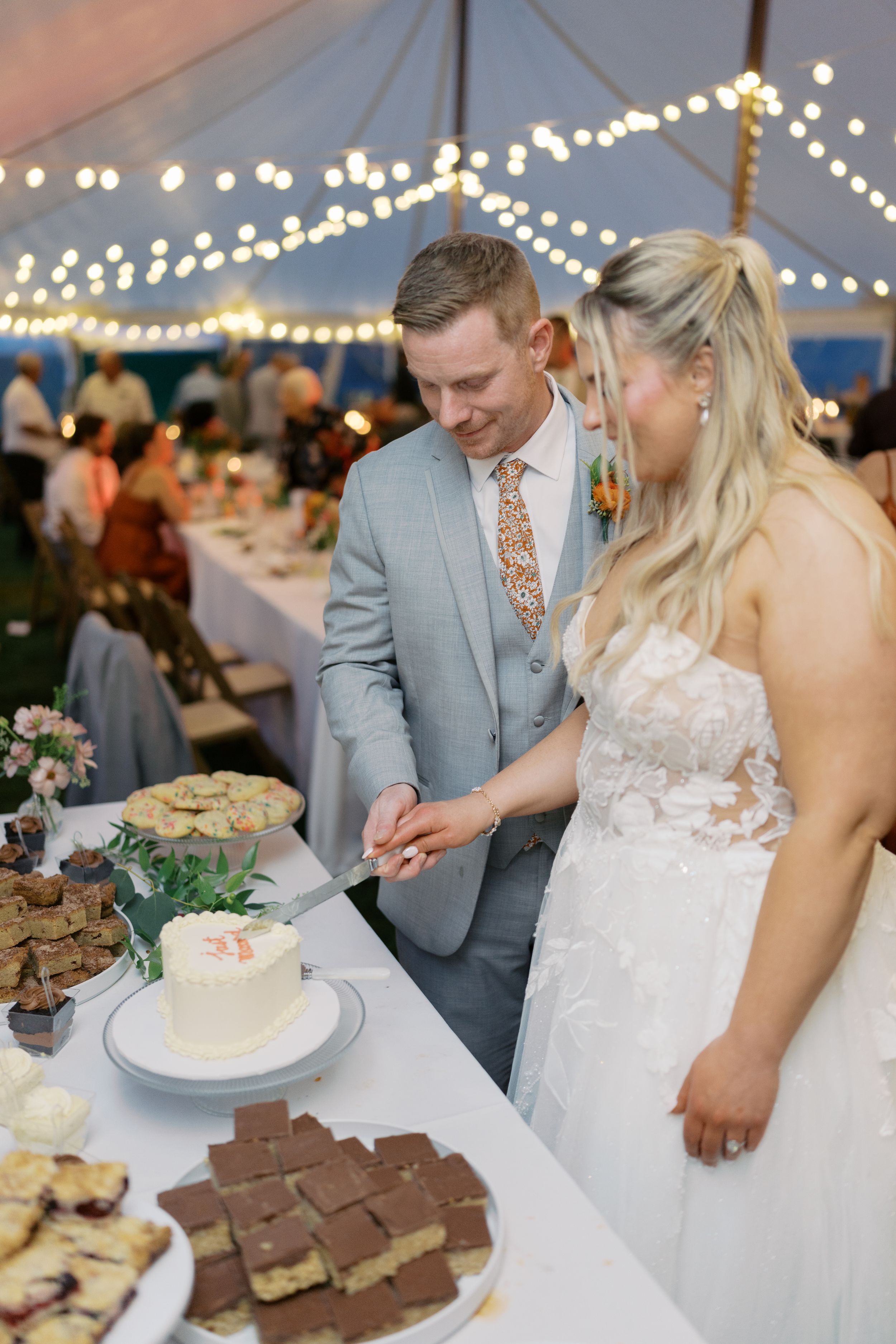 Bride and groom cutting white wedding cake under string lights at elegant tented reception with dessert table.