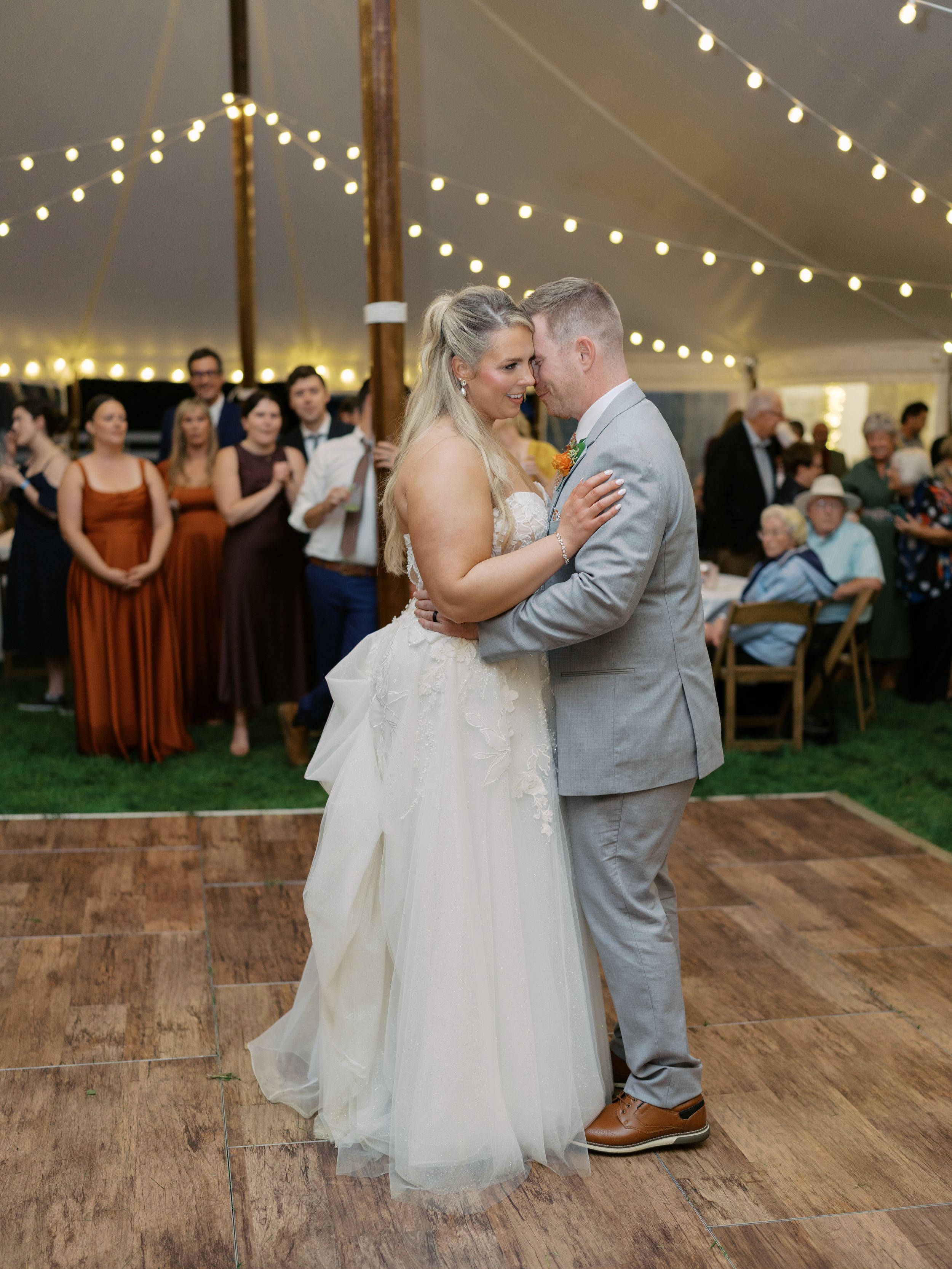 Bride and groom share their first dance under string lights in a white wedding tent surrounded by guests.