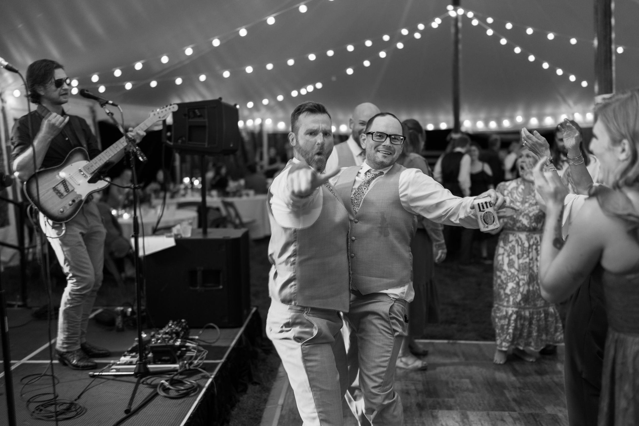 Two men dance joyfully at a wedding reception under string lights with a live band performing on stage.