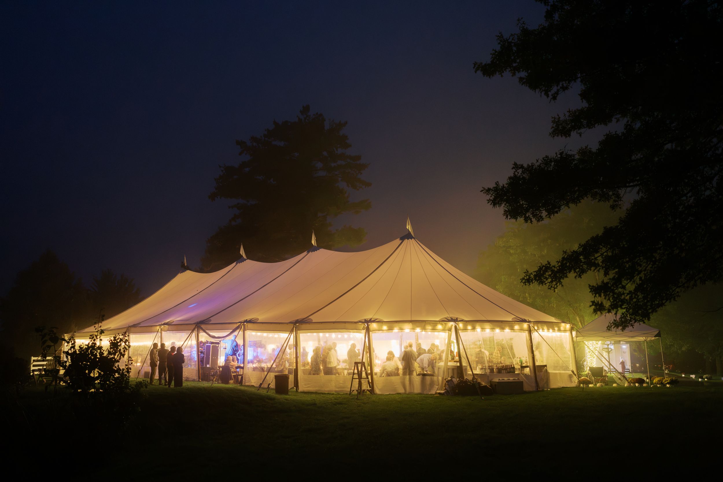 Elegant white wedding tent glowing with warm lights at night, surrounded by trees in a lush outdoor setting.