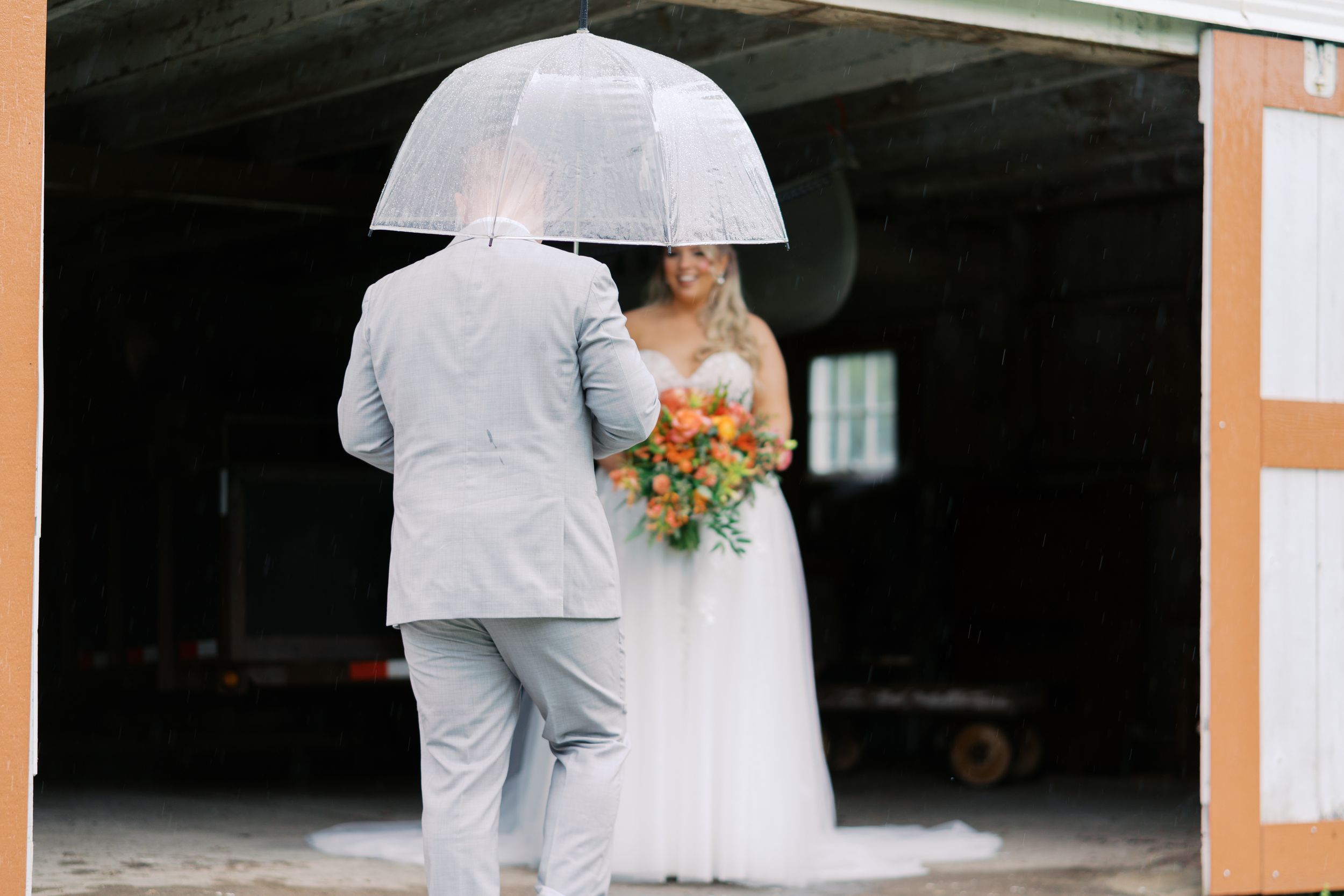 Bride in white gown holding colorful bouquet stands with groom in gray suit under clear umbrella in barn doorway.
