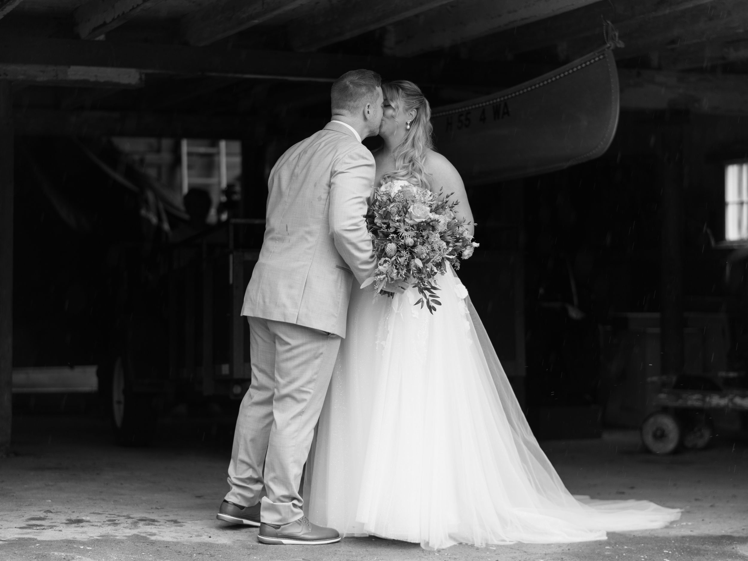 Black and white photo of a bride and groom sharing a kiss, bride holding a bouquet, inside a rustic boathouse.