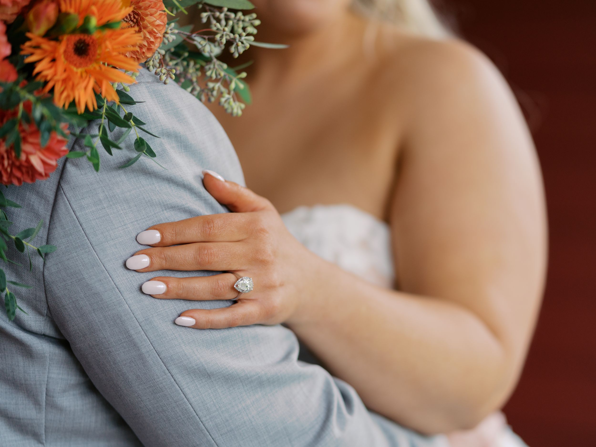 Bride with French manicure and diamond engagement ring embracing groom in gray suit, with orange floral bouquet.