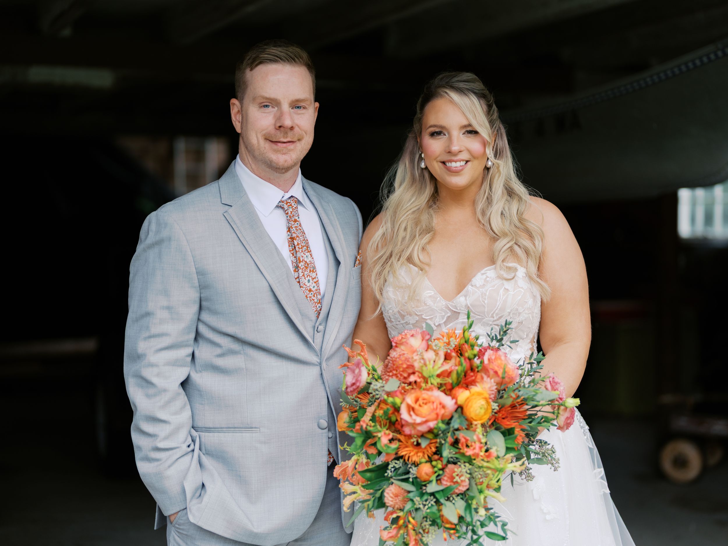 Bride and groom posing together, bride holding vibrant orange bouquet, groom in light gray suit with floral tie.