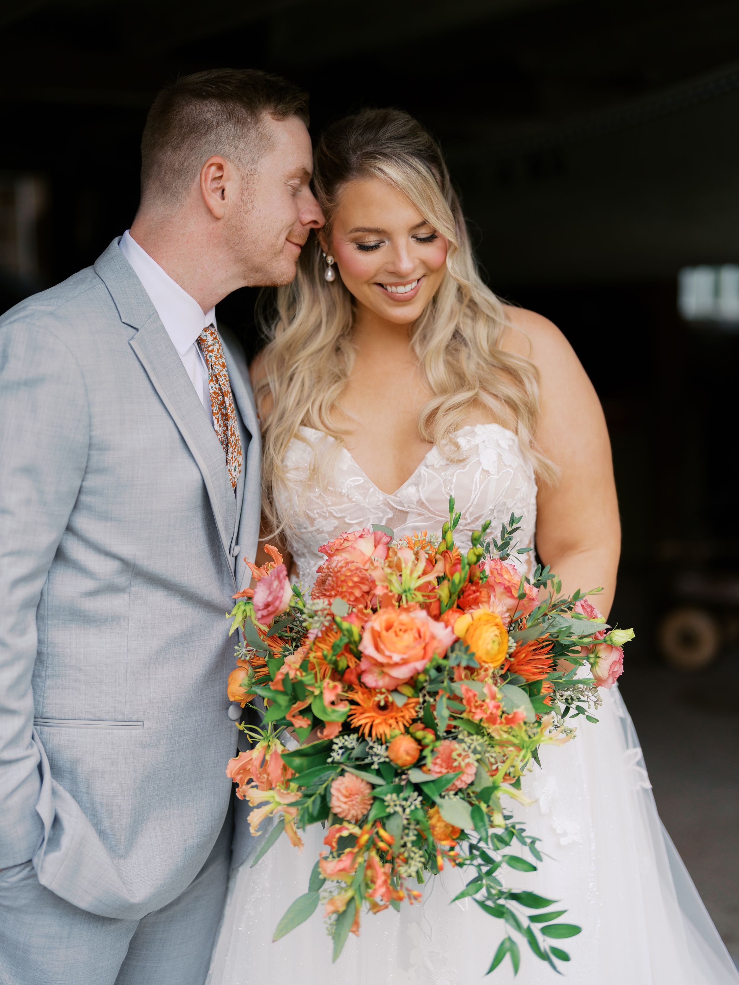 Bride in white lace gown holds vibrant orange bouquet as groom in gray suit whispers in her ear.