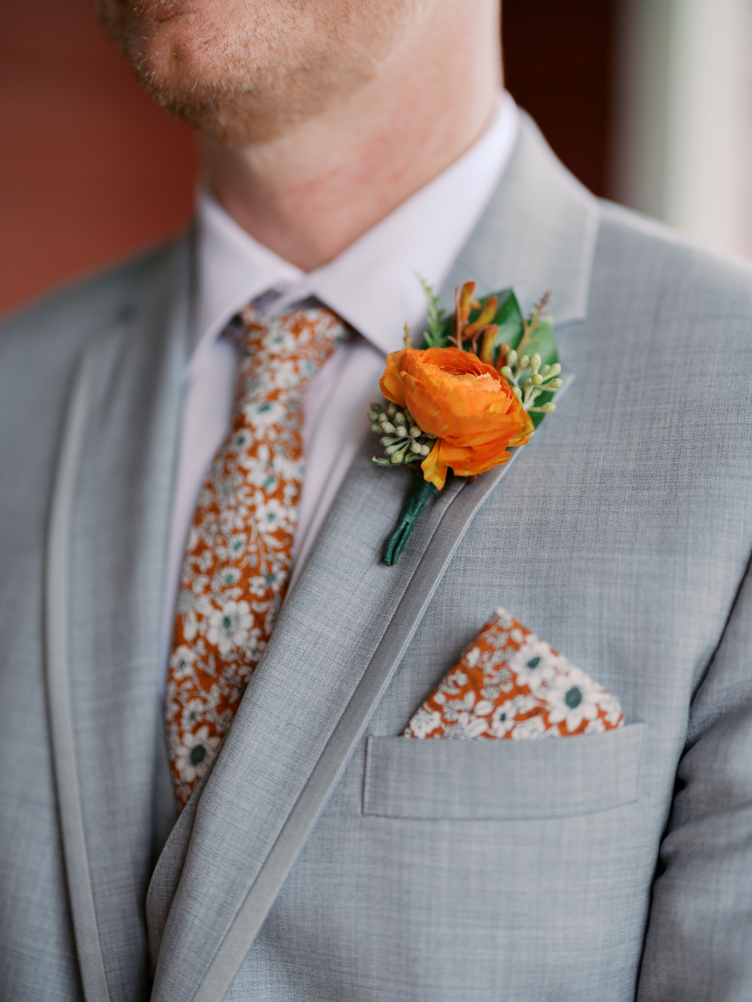 Groom in gray suit with orange ranunculus boutonniere, floral tie and matching pocket square at wedding.