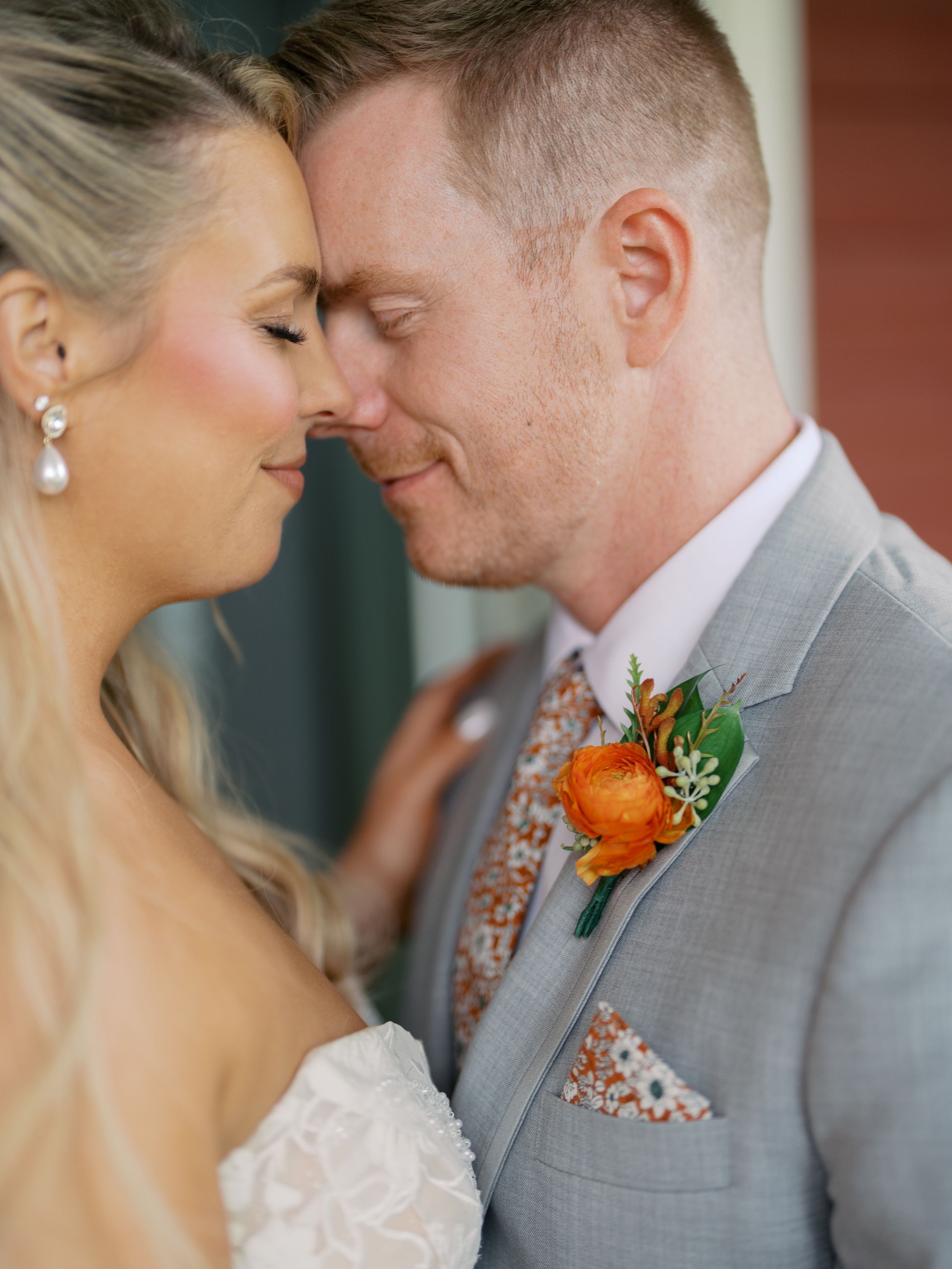 A bride and groom share an intimate moment, touching foreheads, groom wearing a gray suit with orange boutonniere.