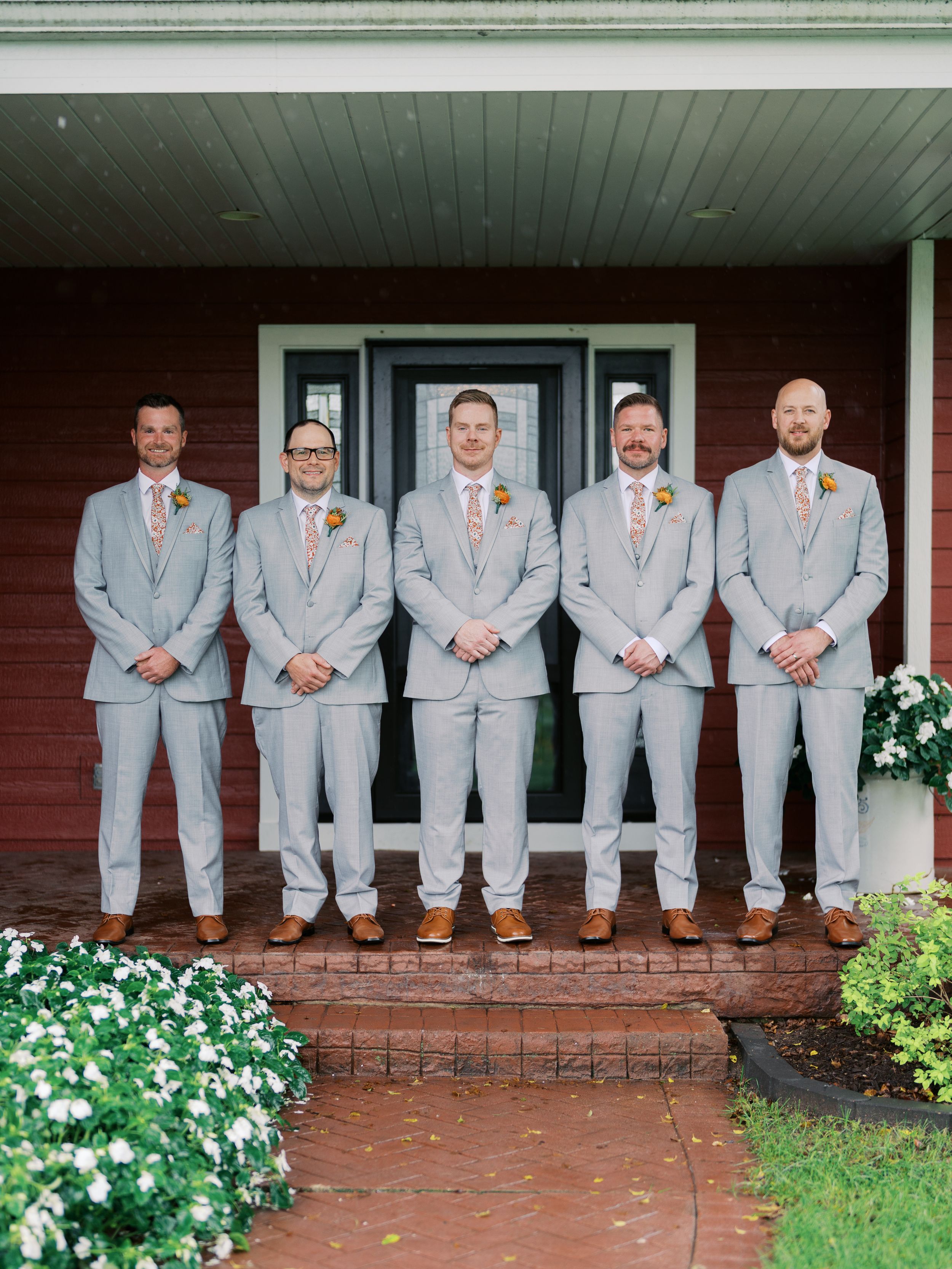 Five groomsmen in matching light gray suits with boutonnieres pose on brick steps outside a red house.