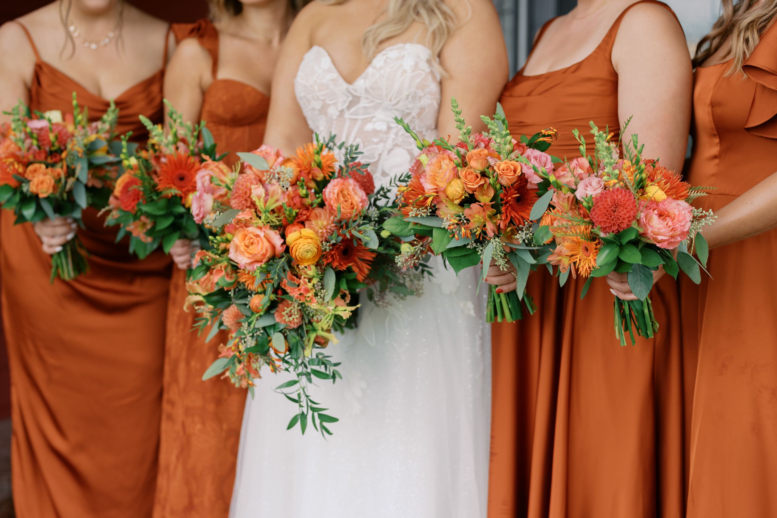 Bride in white lace gown and bridesmaids in rust orange dresses holding vibrant orange and pink floral bouquets.