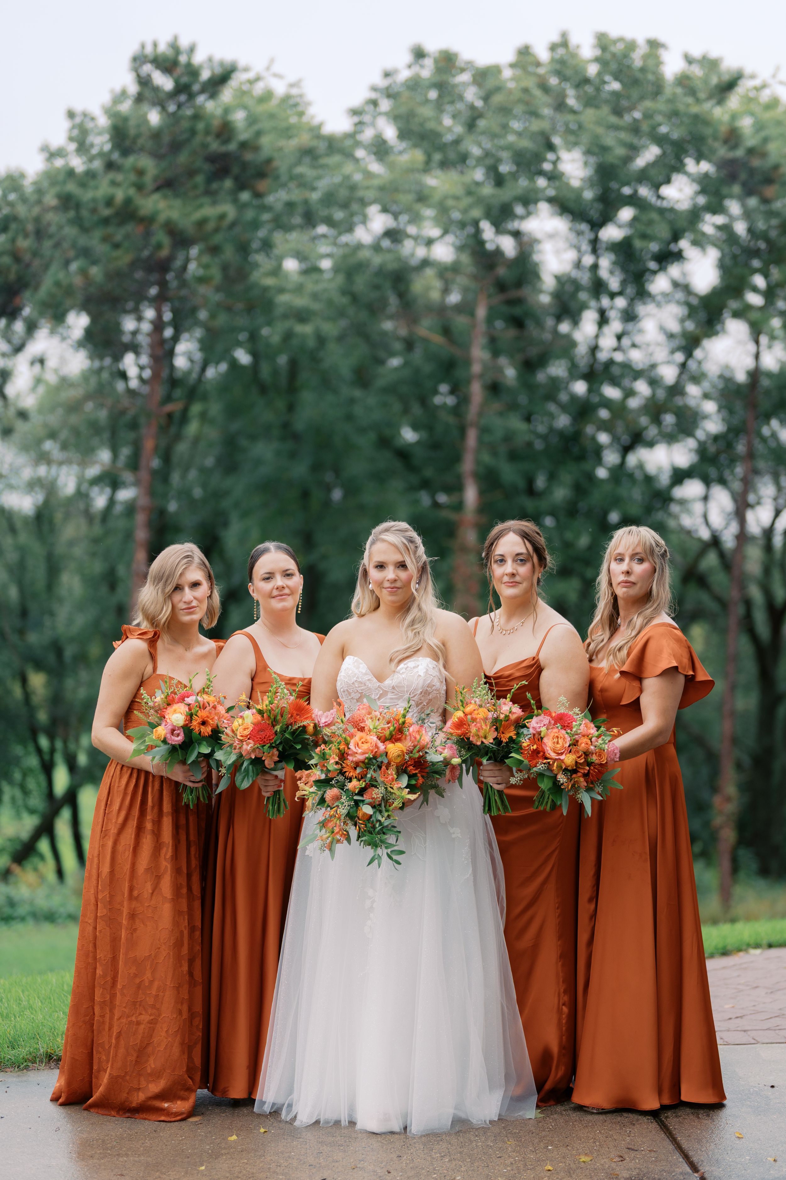 Bride in white gown with four bridesmaids in rust orange satin dresses holding bouquets outdoors.