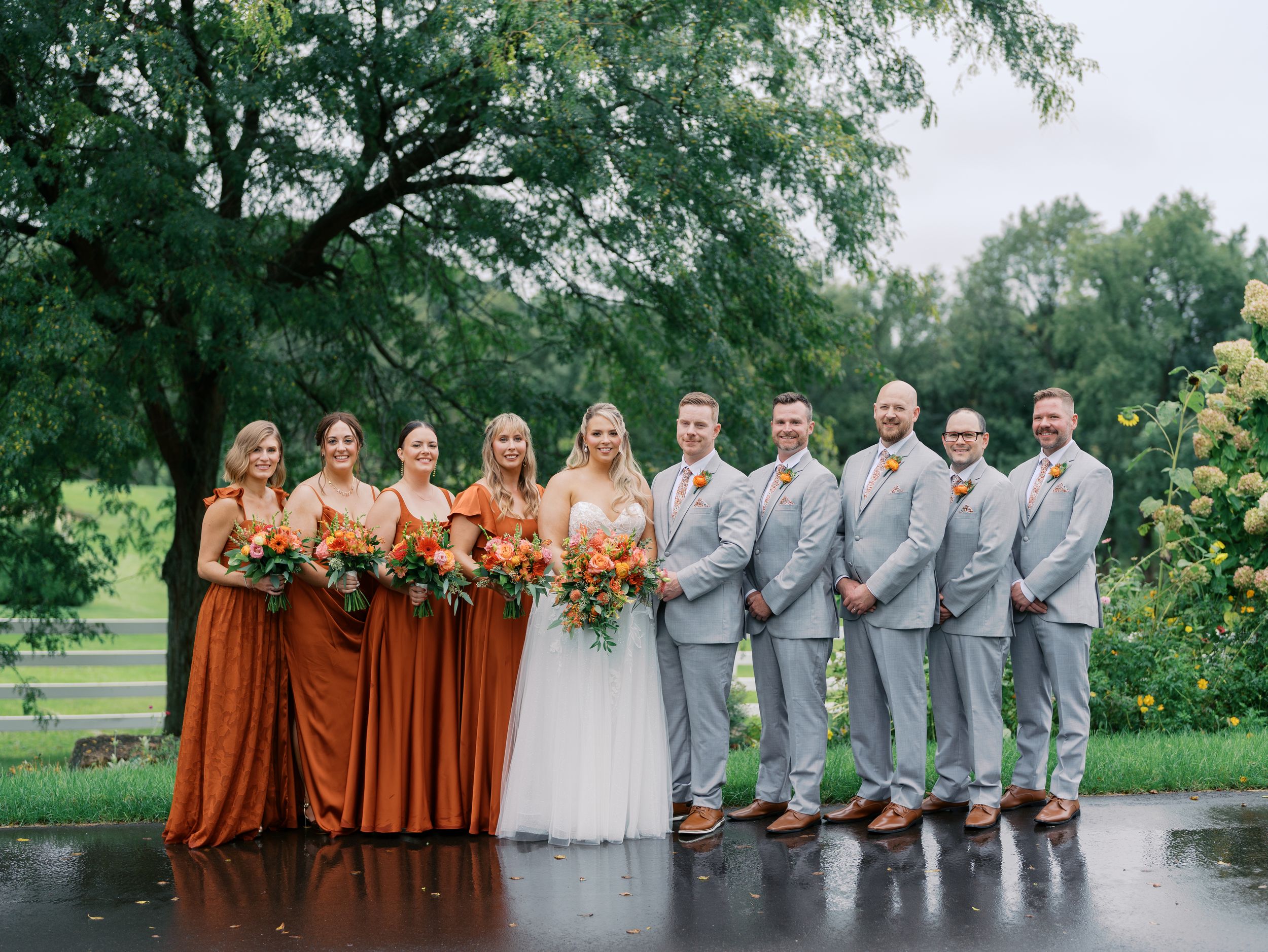 Wedding party poses outdoors in rust bridesmaid gowns and gray suits with fall floral bouquets on a rainy day.