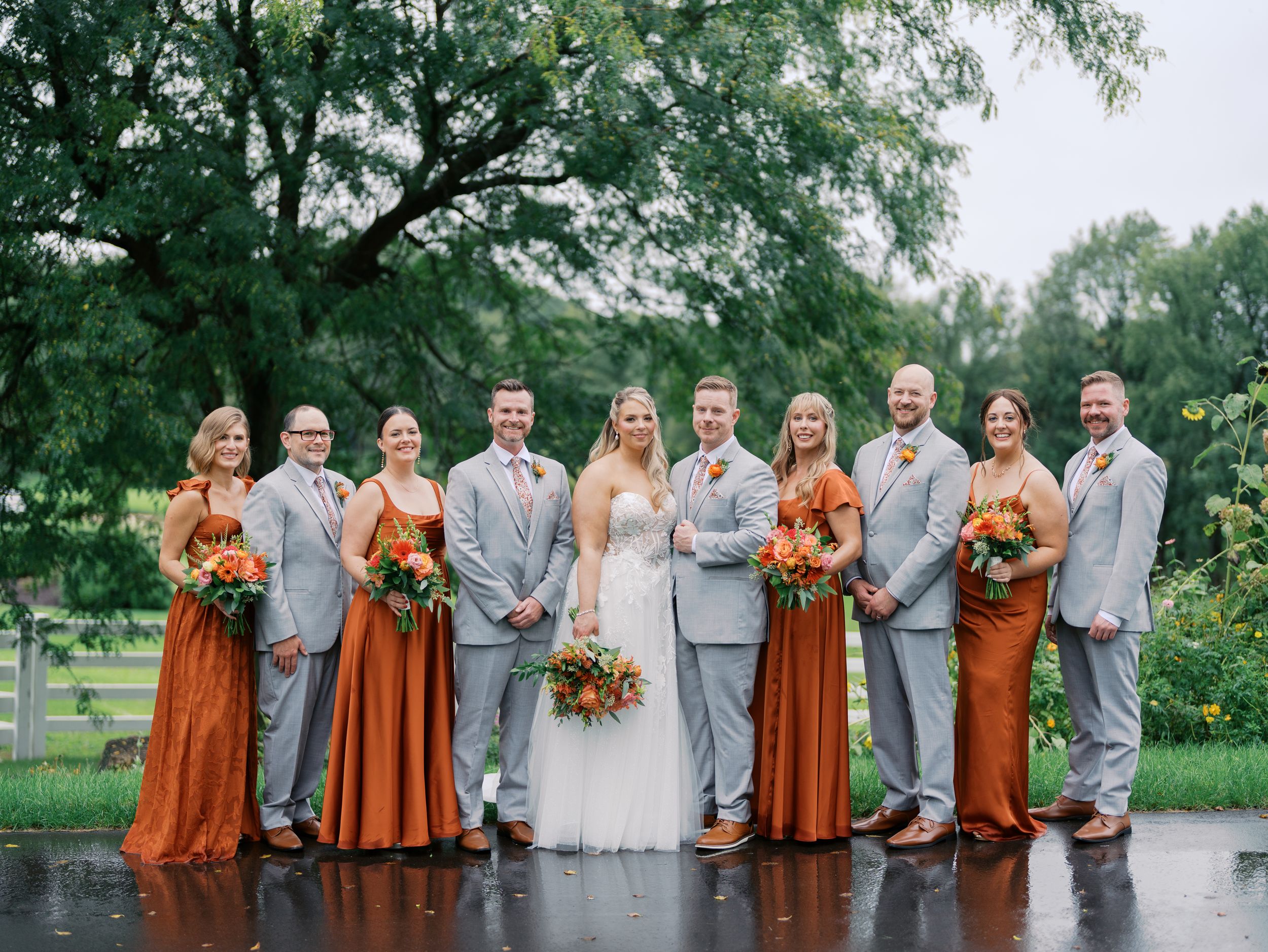 Wedding party in gray suits and rust orange bridesmaid dresses posing outdoors on a rainy day.