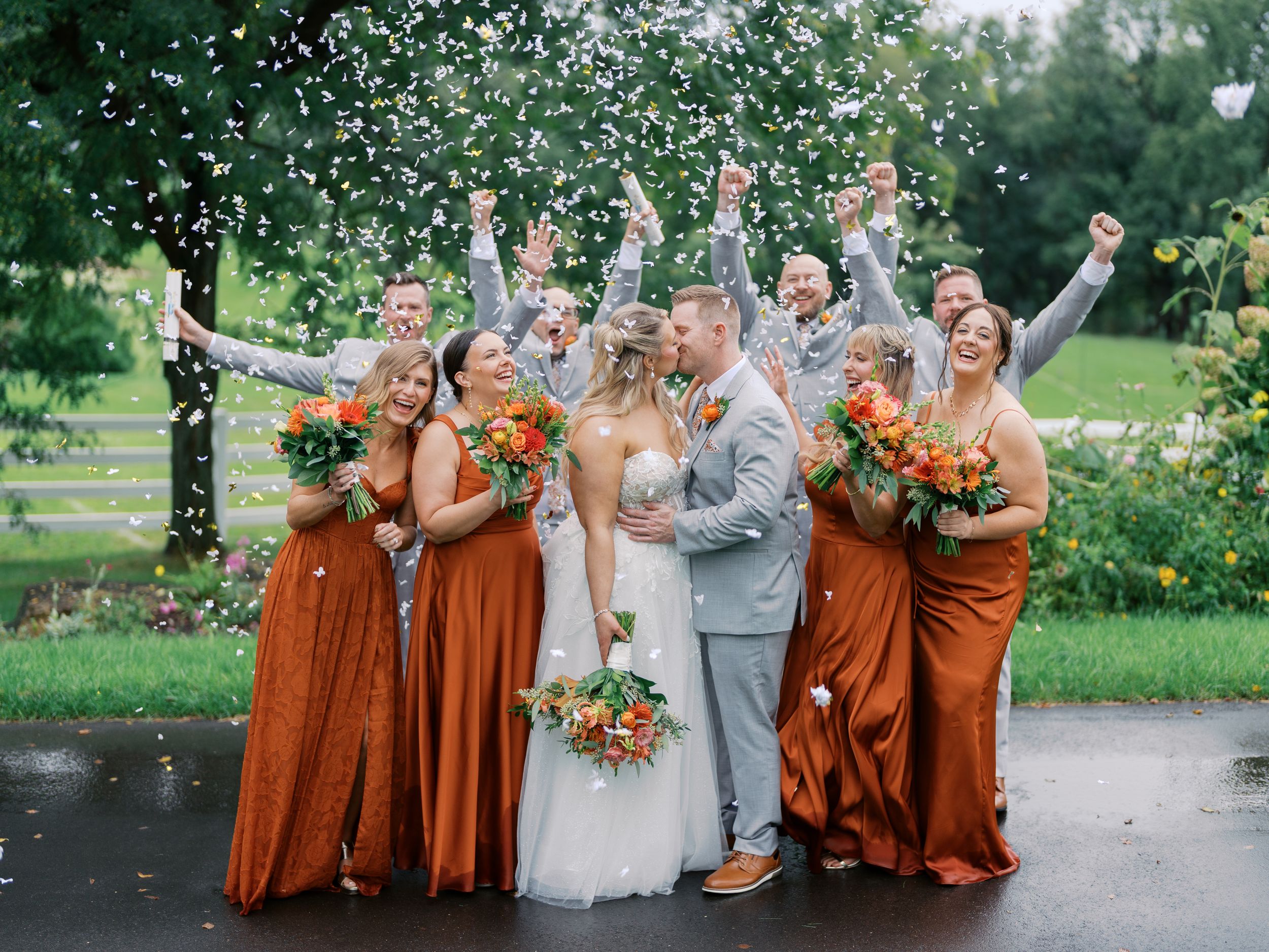 Wedding party celebrates with confetti toss, bridesmaids in rust orange gowns flanking bride and groom outdoors.