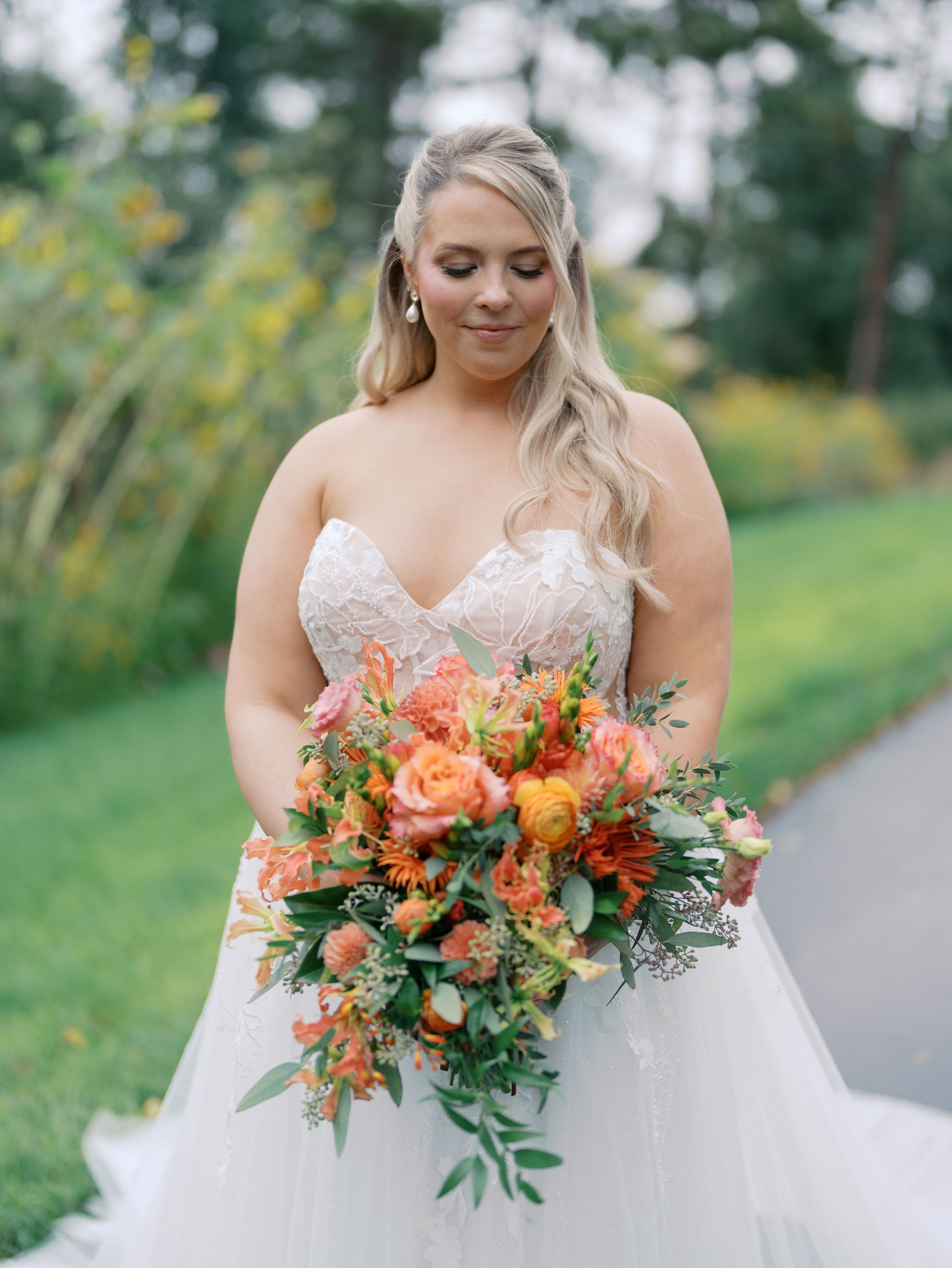 Bride in strapless lace ballgown holding vibrant orange and pink wedding bouquet with greenery outdoors.