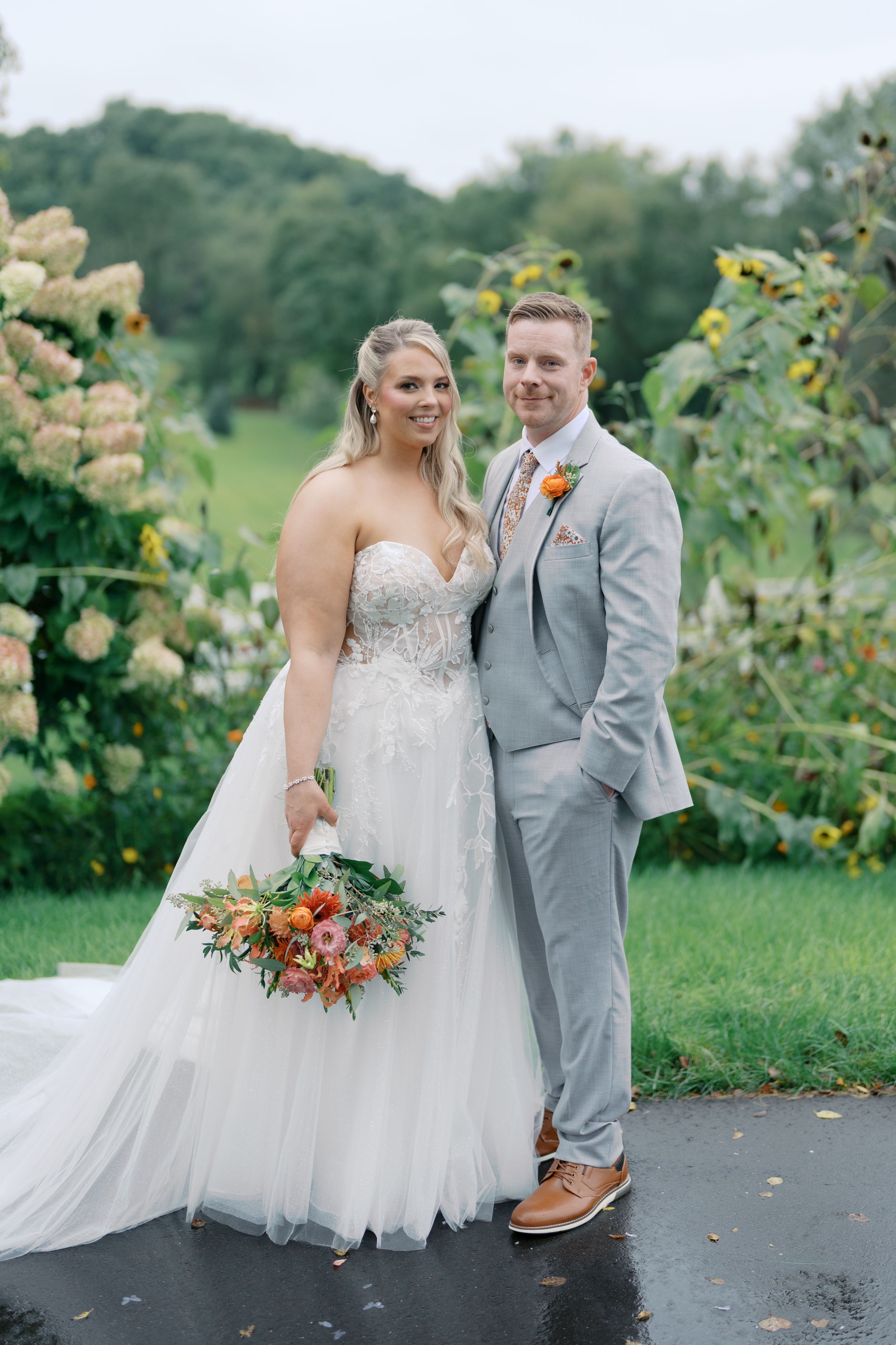 Bride in white gown holding bouquet stands with groom in gray suit outdoors near sunflowers and hydrangeas.