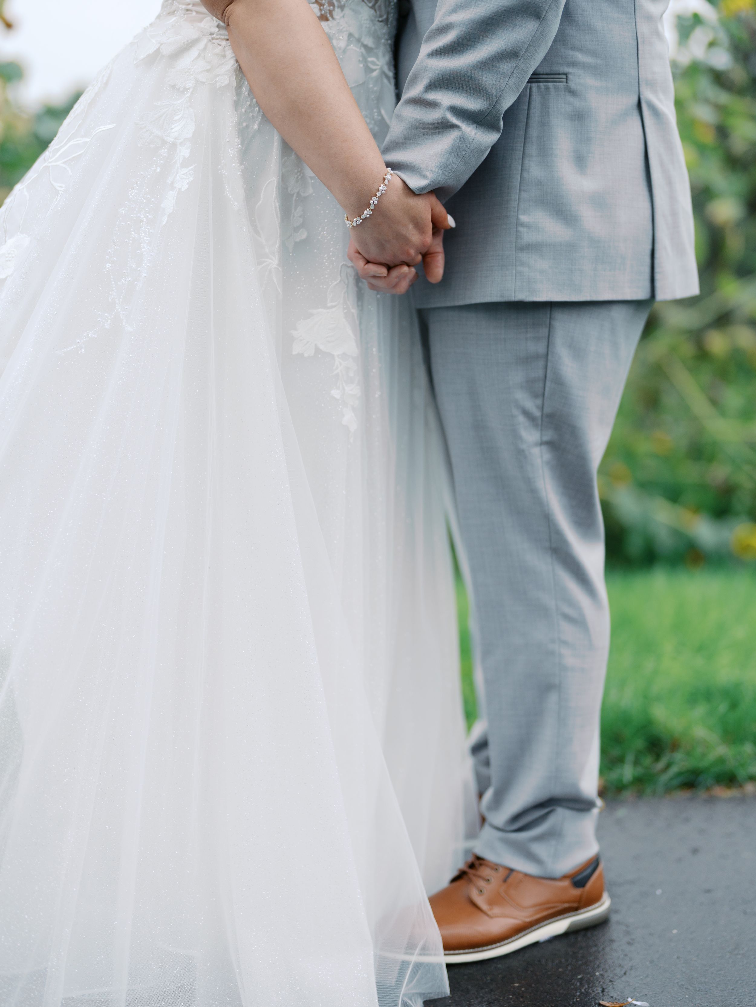 A bride in a white lace gown and groom in a gray suit hold hands tenderly during their outdoor wedding ceremony.