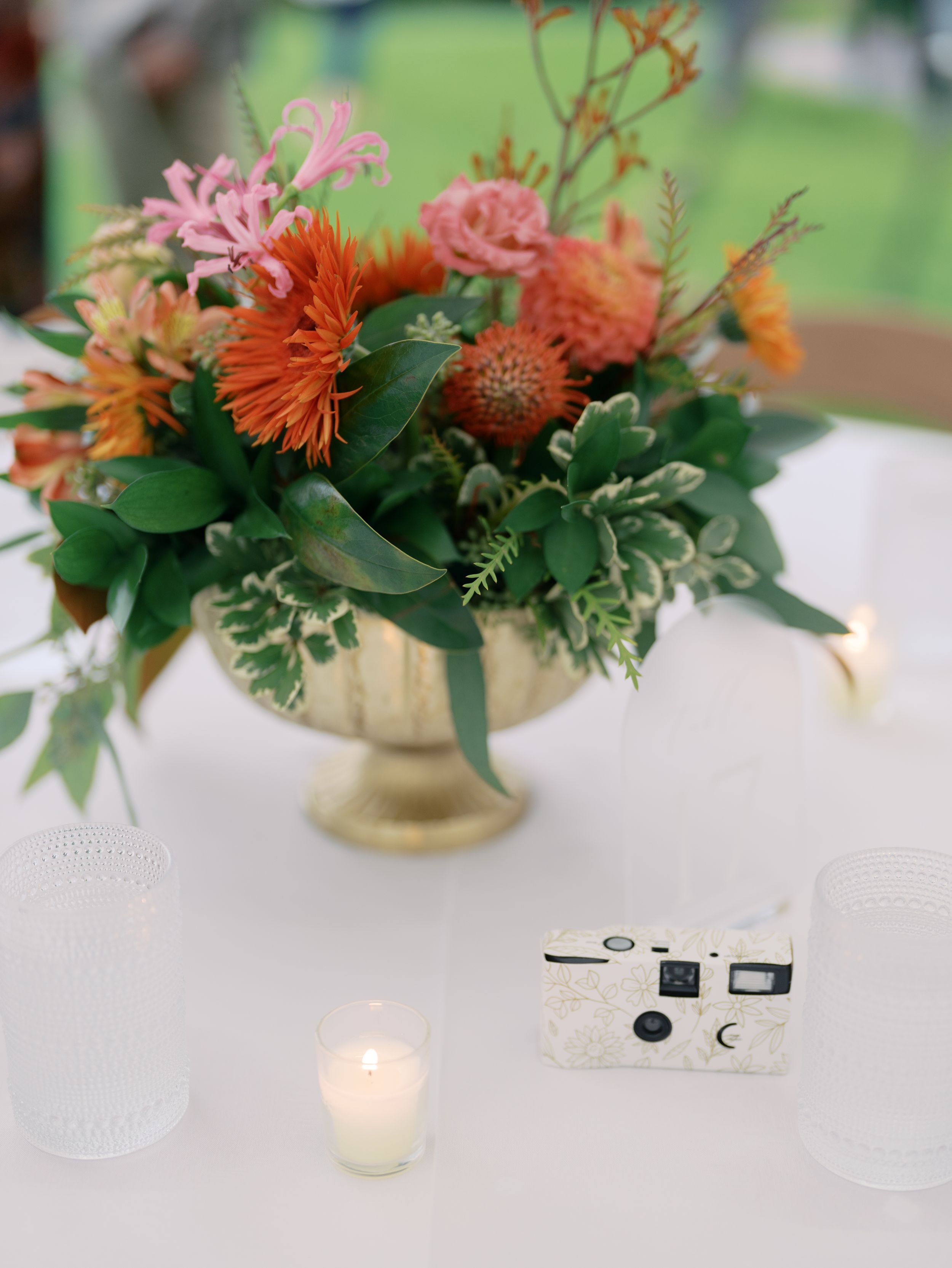 Gold bowl centerpiece with orange and pink flowers, greenery, candles, and a disposable camera on a white tablecloth.