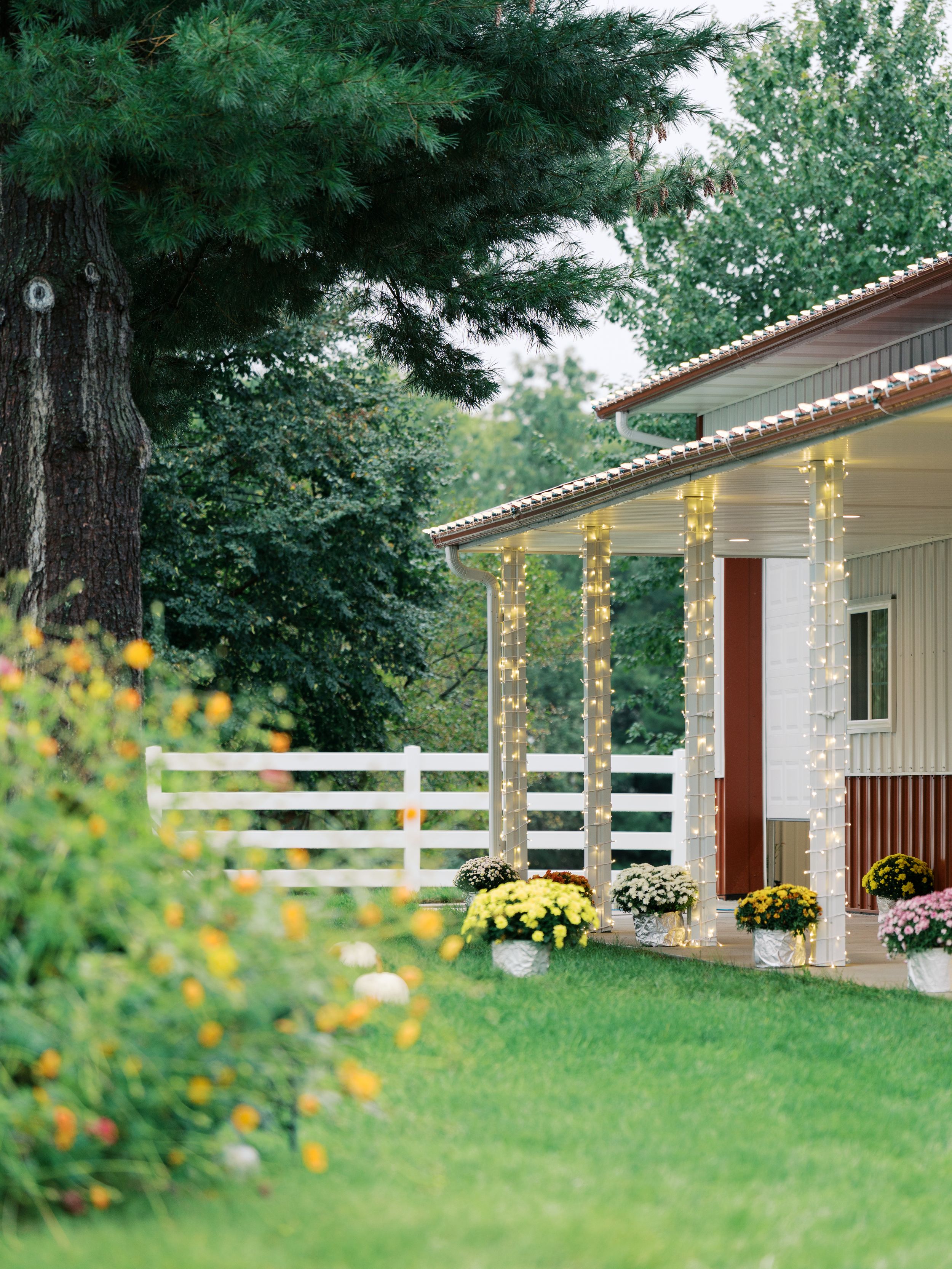 Charming farmhouse porch with string lights on columns, white fence, colorful mums, and lush green lawn at dusk.