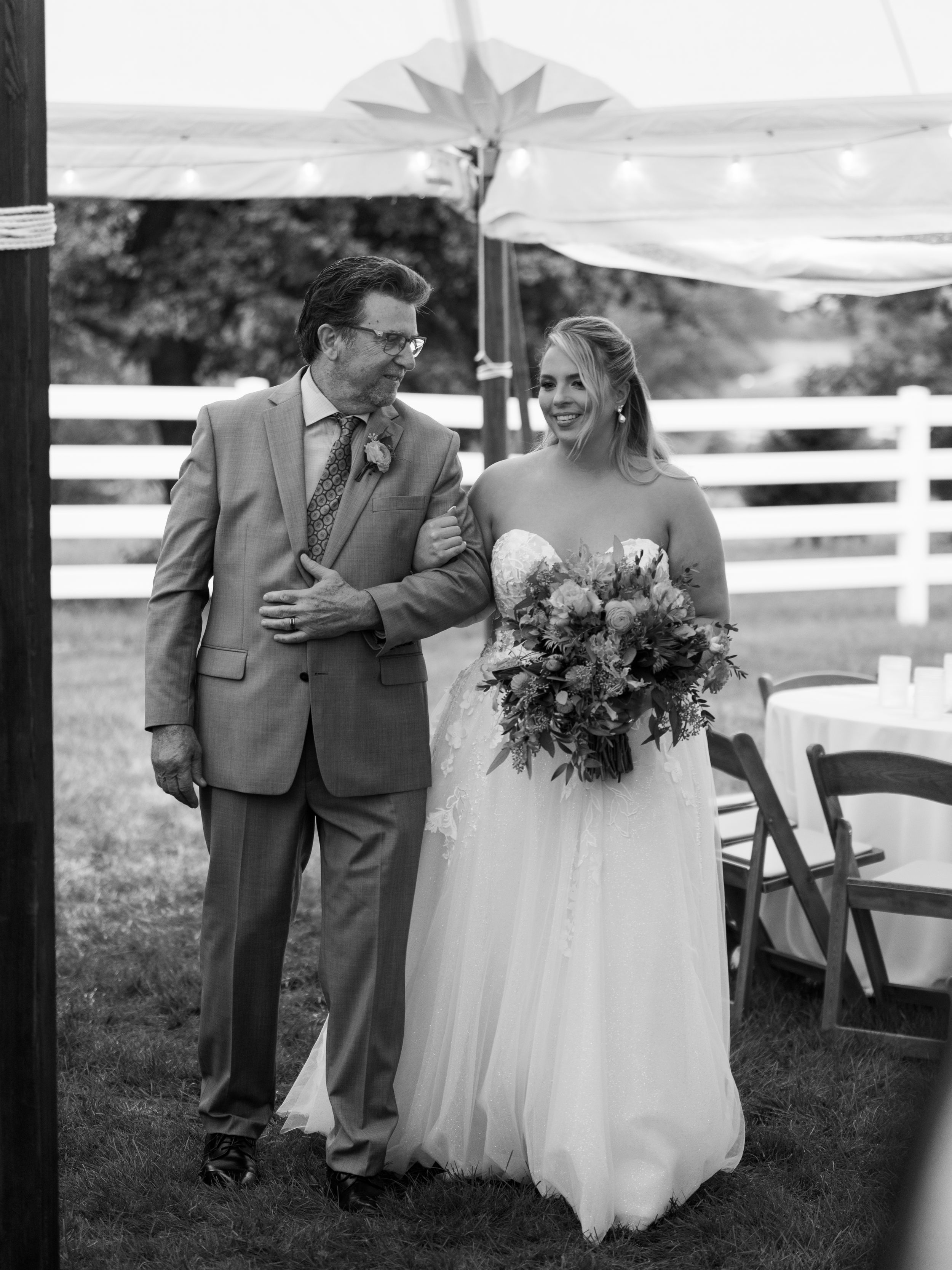 Black and white photo of a smiling bride in a white gown holding a bouquet, walking with a man in a suit at an outdoor wedding.