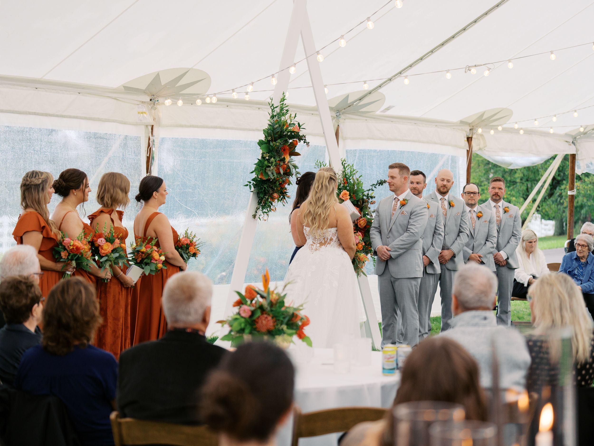 Outdoor wedding ceremony under a white tent with bridesmaids in orange dresses and groomsmen in gray suits at floral arch.
