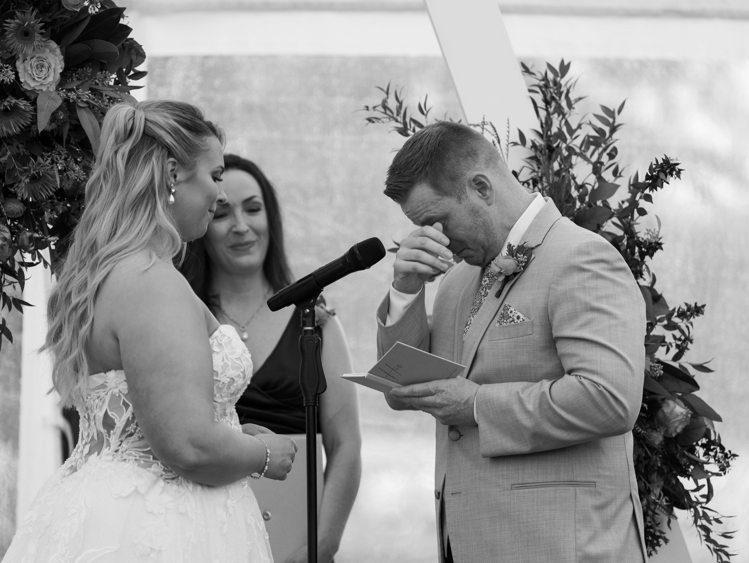 Groom reads emotional wedding vows to bride at outdoor ceremony while officiant watches in black and white photo.