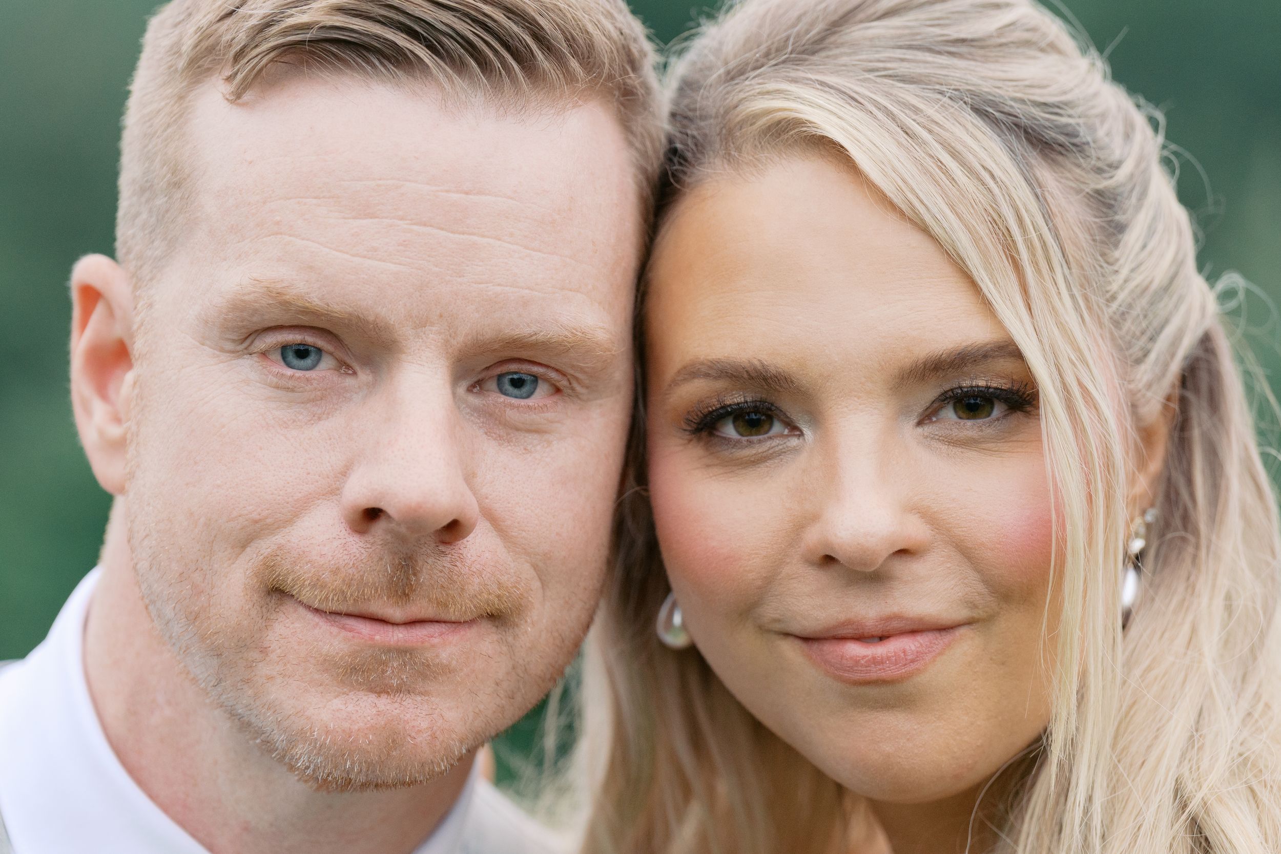 Close-up portrait of a smiling couple, man with red hair and woman with blonde hair, posing outdoors.