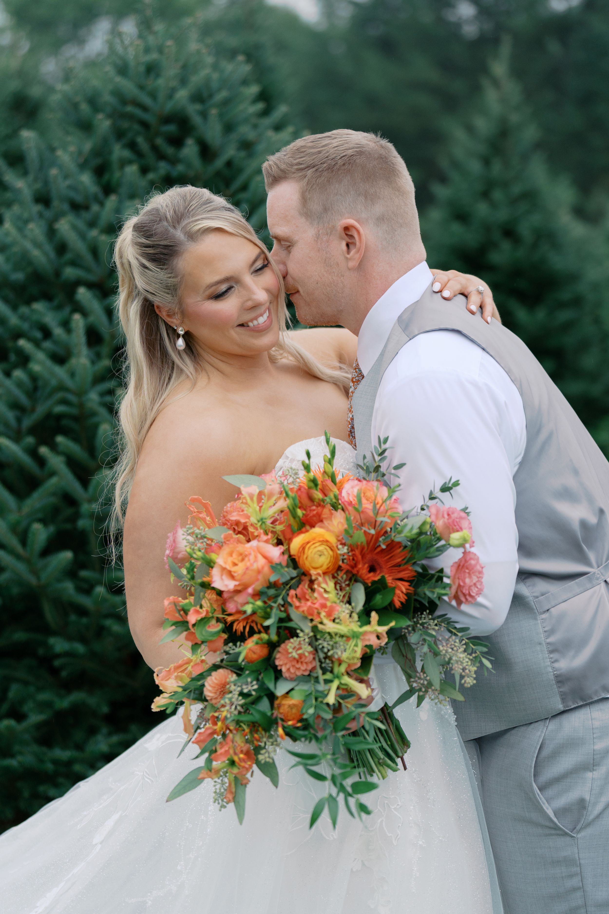 Bride smiling holding orange floral bouquet while groom kisses her cheek outdoors near evergreen trees.