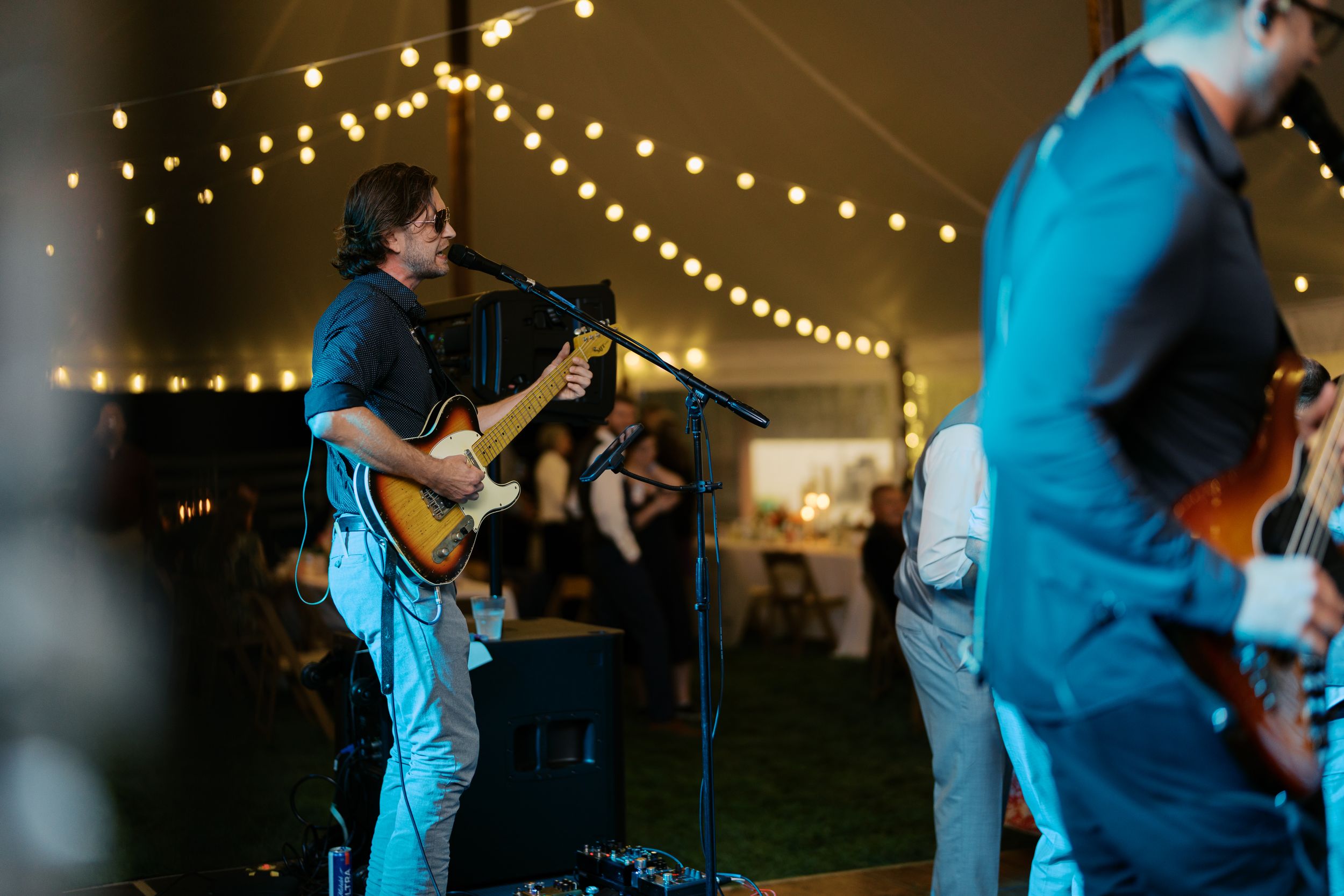 Live band performing under string lights at an outdoor tent wedding reception with guitarist at microphone.