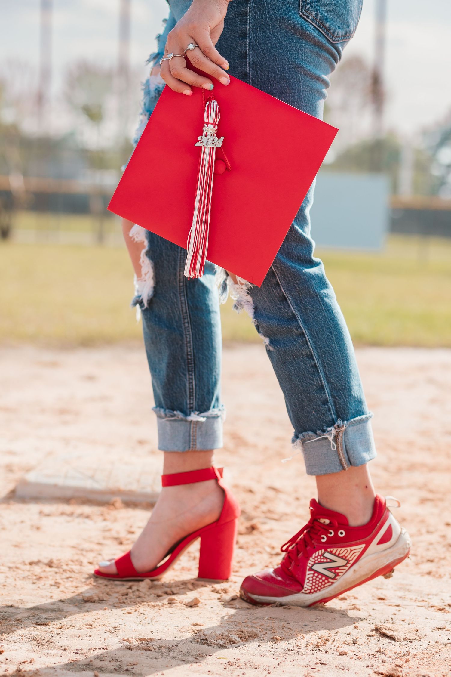 Graduate holding red cap with tassel, wearing mismatched red heels and sneaker with ripped jeans on sandy ground.