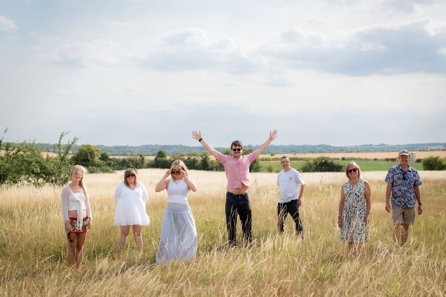 Group of seven people posing in a golden field under cloudy skies, one person raising arms joyfully.