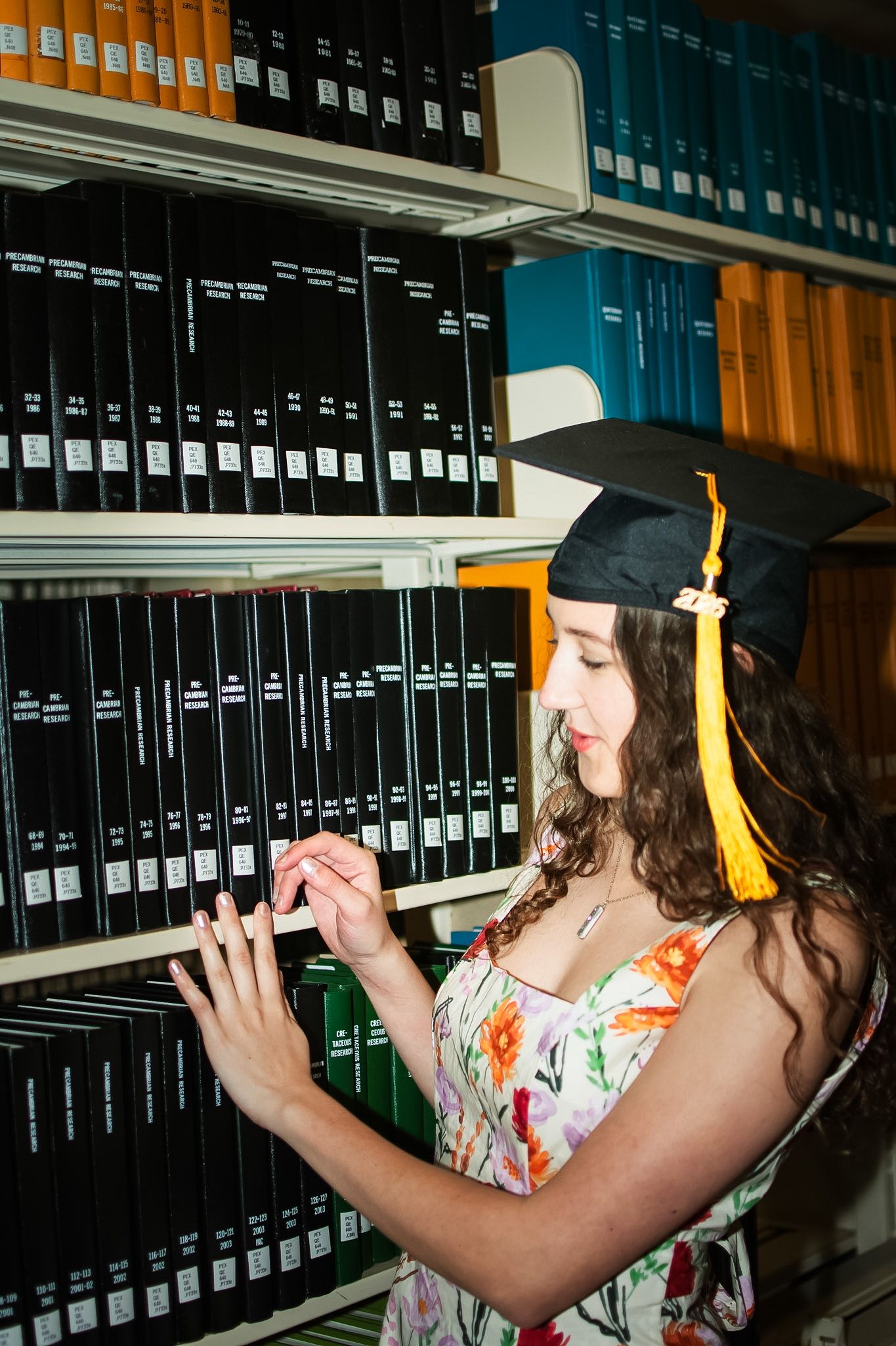Young woman in graduation cap and floral dress browsing books on library shelves.