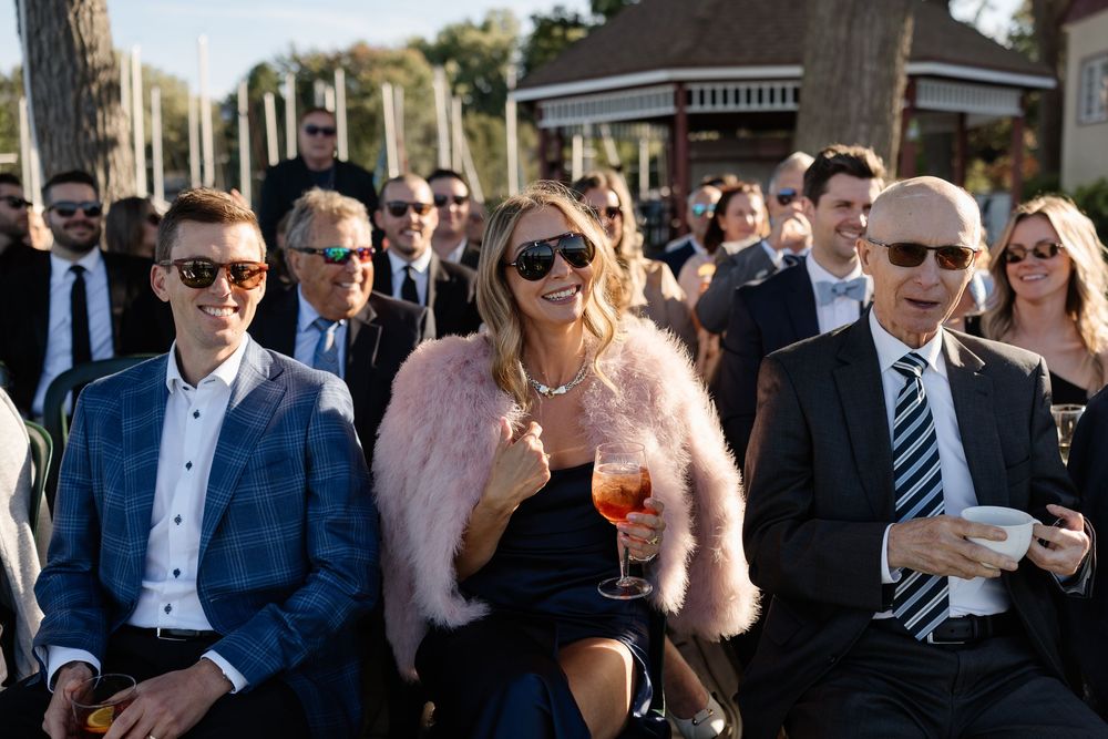 Wedding guests in formal attire seated outdoors, woman in pink fur wrap holding wine glass, smiling crowd.